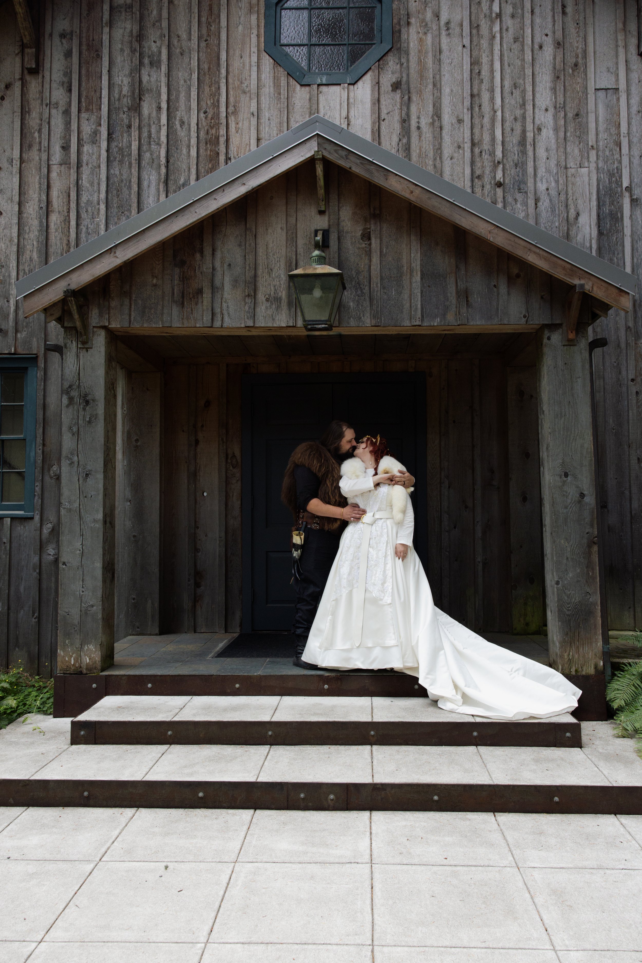 A couple dressed in wedding attire sharing a kiss outside a rustic wooden building with a black door, a lantern hanging above, and a small octagonal window.