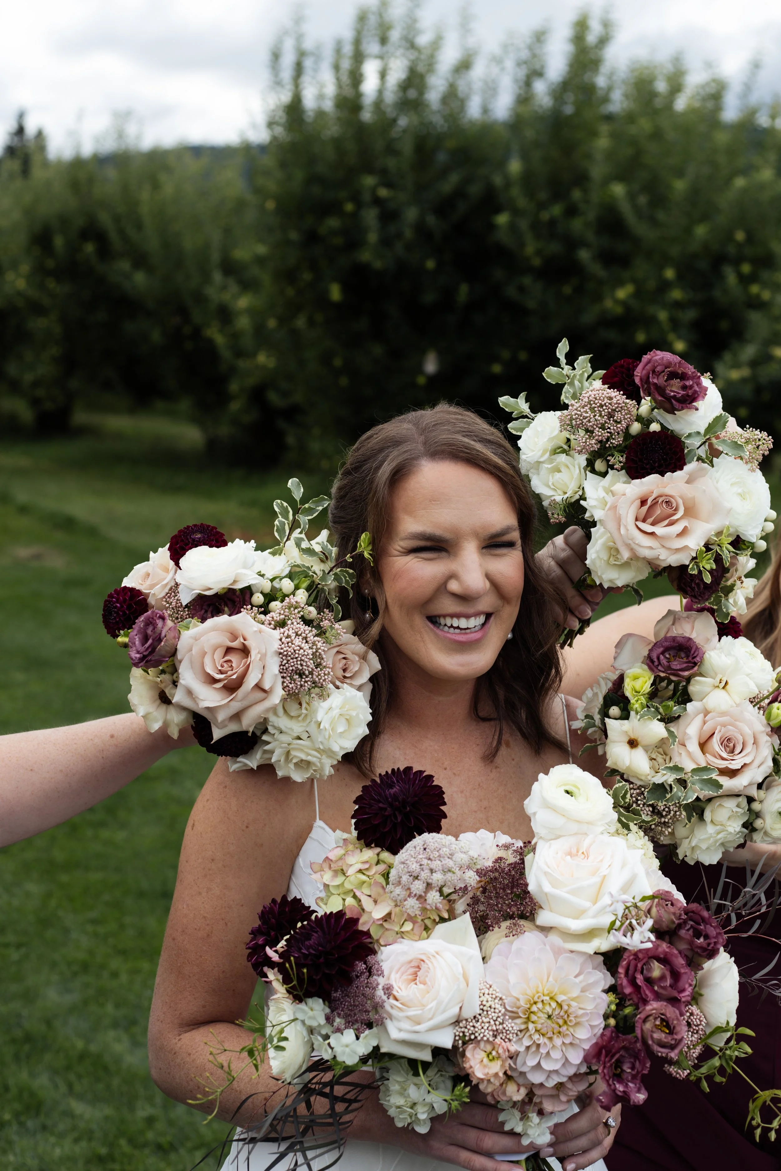 A smiling woman holding a large bouquet of various flowers, including roses and other blooms, outdoors with green trees in the background.