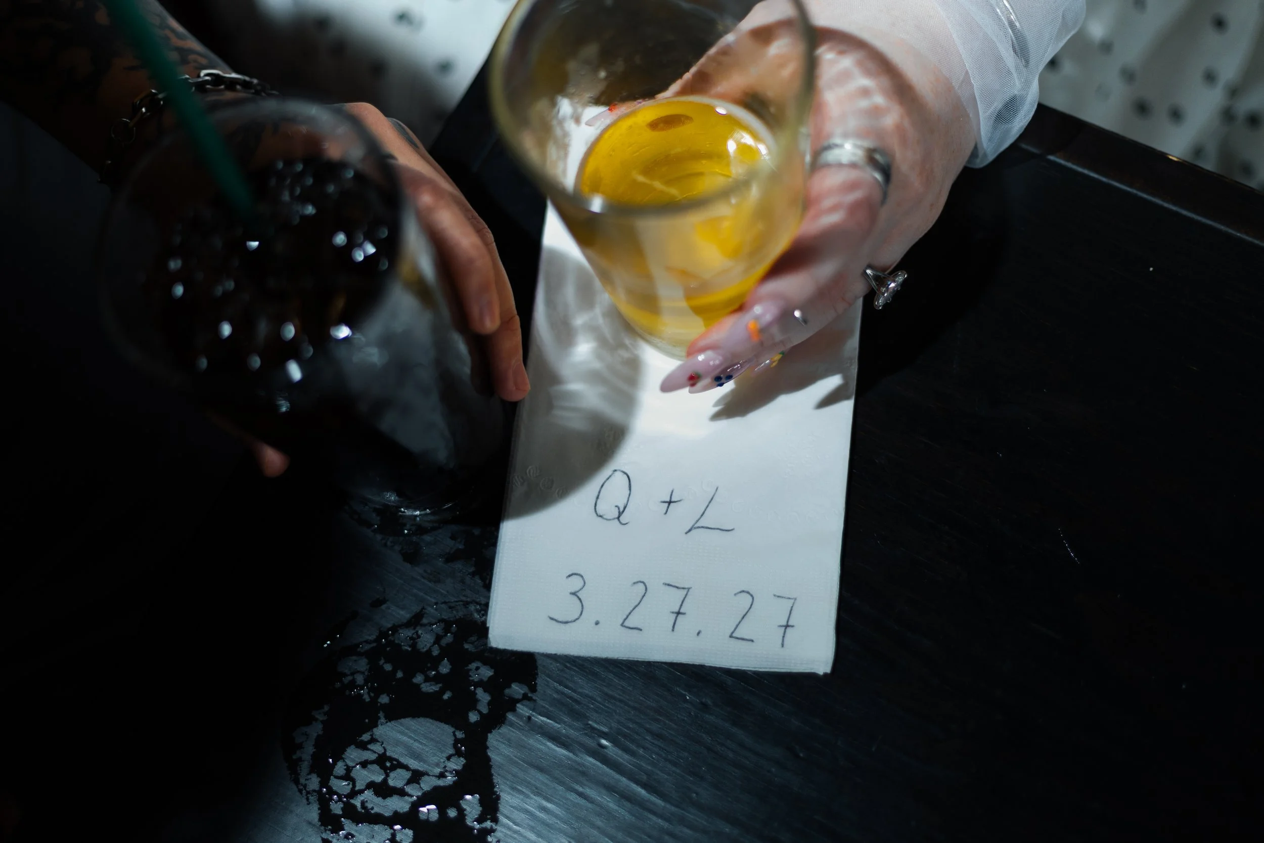 A person holding a glass of what appears to be beer or a similar beverage, with notes and a handwritten note showing a price of 3.27 dollars on a white paper. The background is dark with some objects, including a beer bottle and a black lace item.