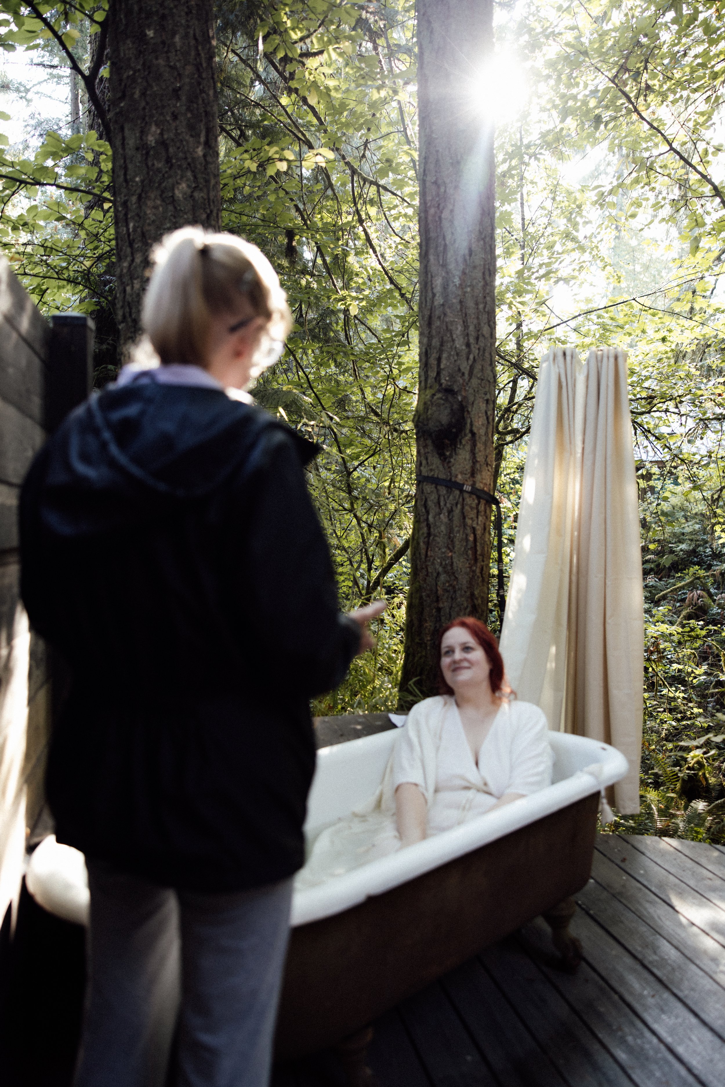 A woman in a white dress sitting in a bathtub outdoors amidst trees, with sunlight filtering through the leaves; a person standing nearby, slightly out of focus, holding a phone.