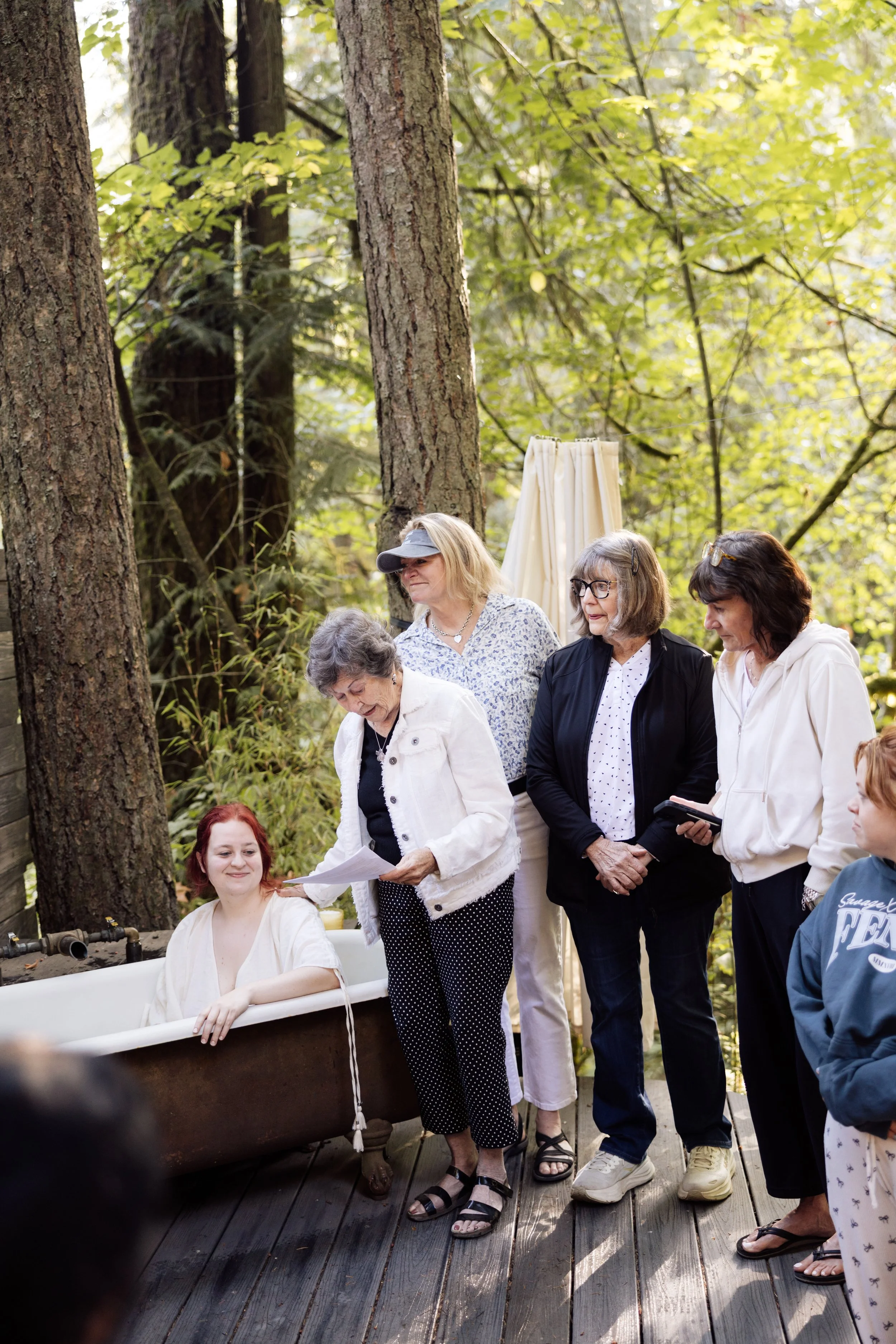 A group of women, mostly elderly, are gathered outdoors in a wooded area. Some are standing, and one woman is sitting in a vintage bathtub. The women are engaged in an activity, with one woman reading from papers. The scene appears casual and social,