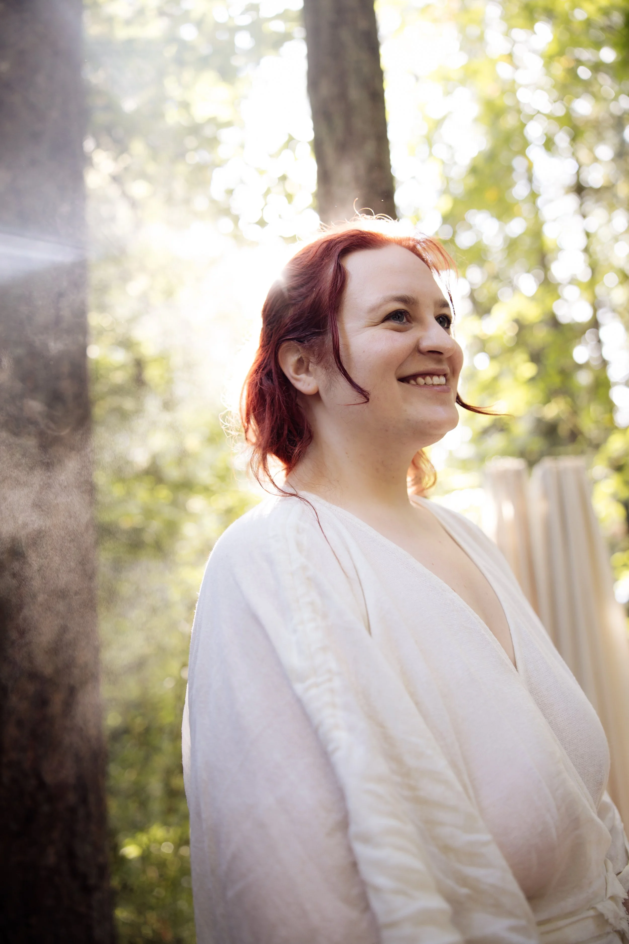 A woman with red hair smiling outdoors in sunlight, surrounded by trees and greenery.