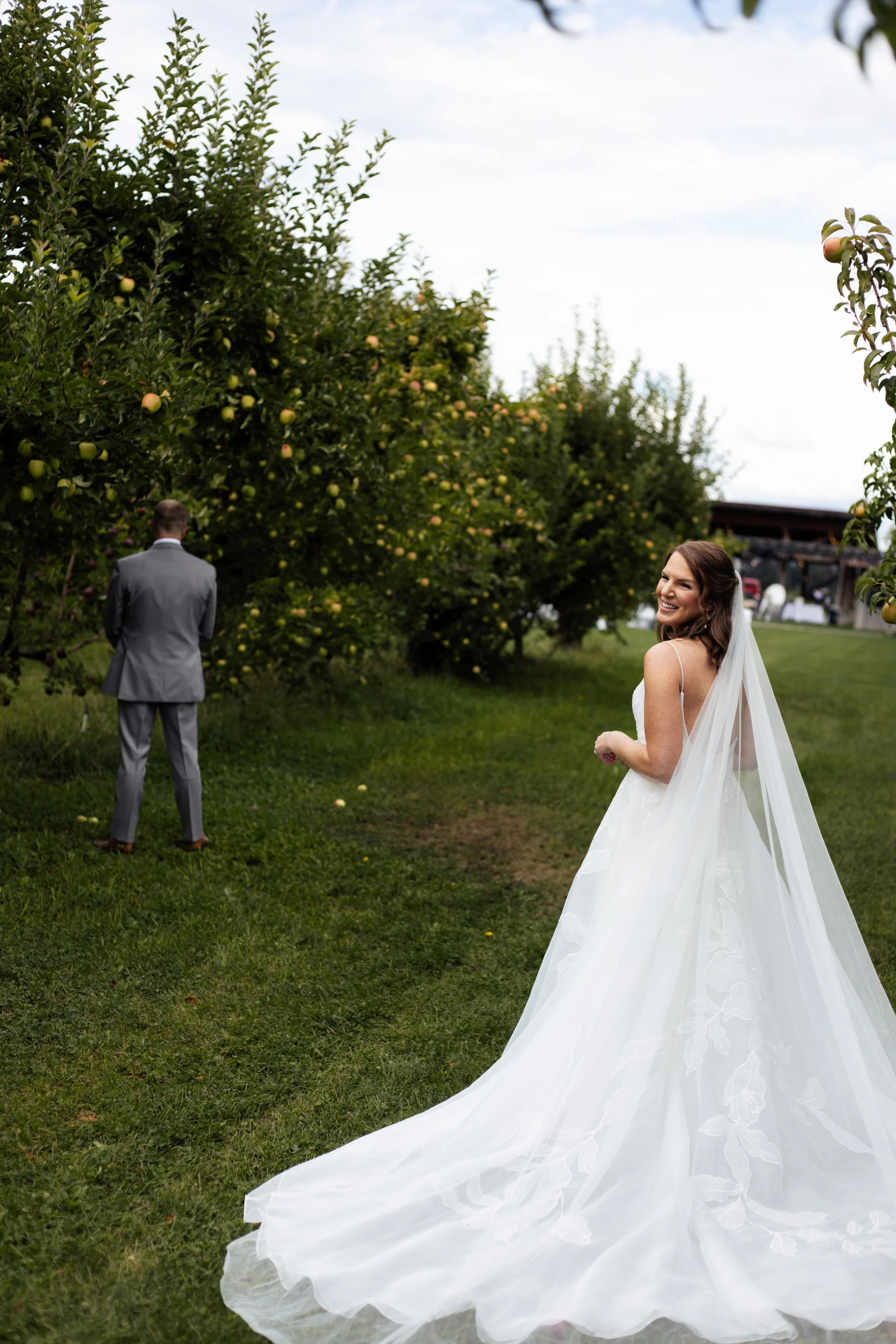 A bride in a white wedding dress smiling and looking back, with a groom in a gray suit standing near apple trees. The scene is outdoors on a grassy area.