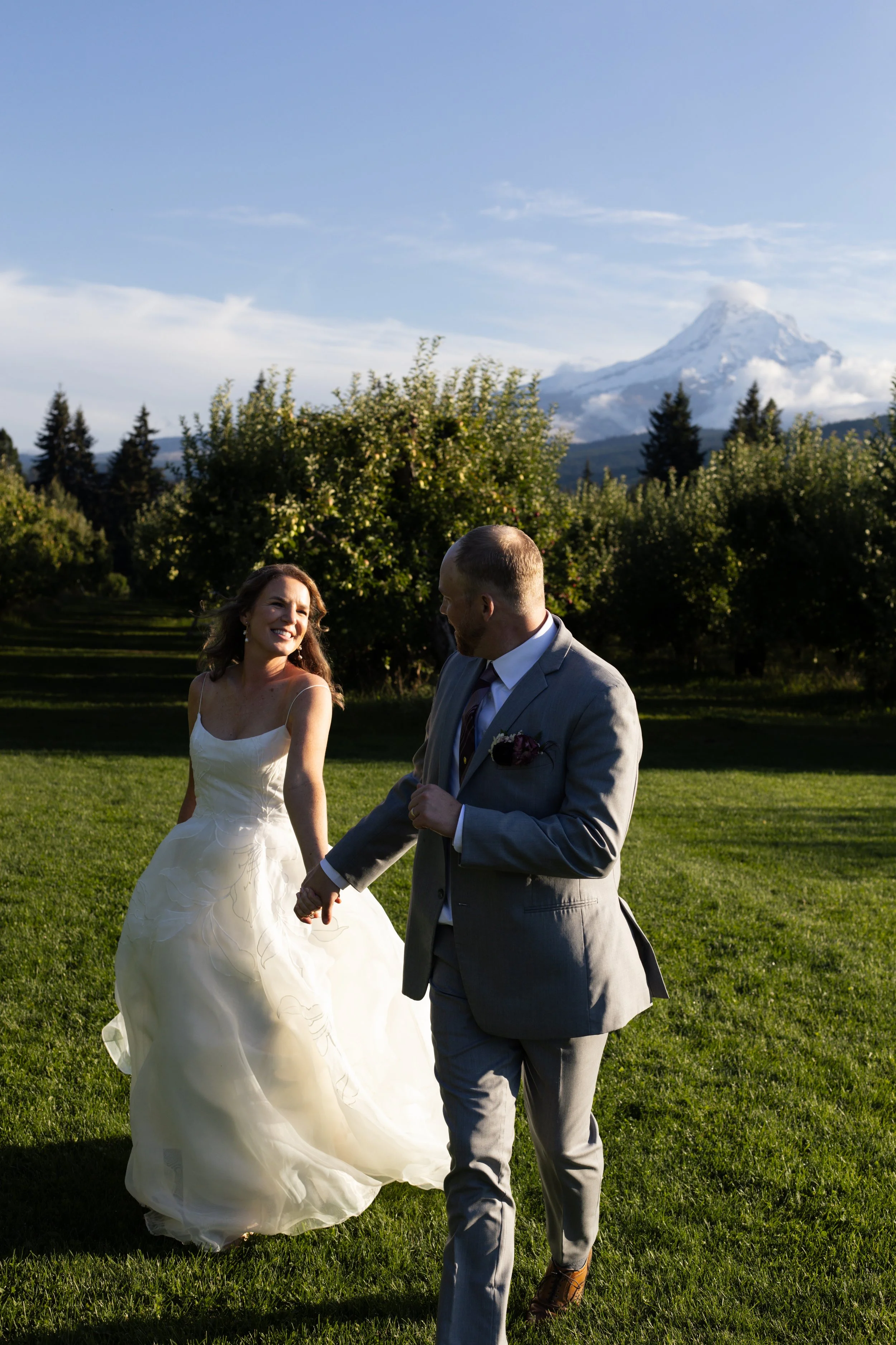 A bride and groom holding hands outdoors with a snow-capped mountain in the background, green grass and trees around, during a wedding celebration.