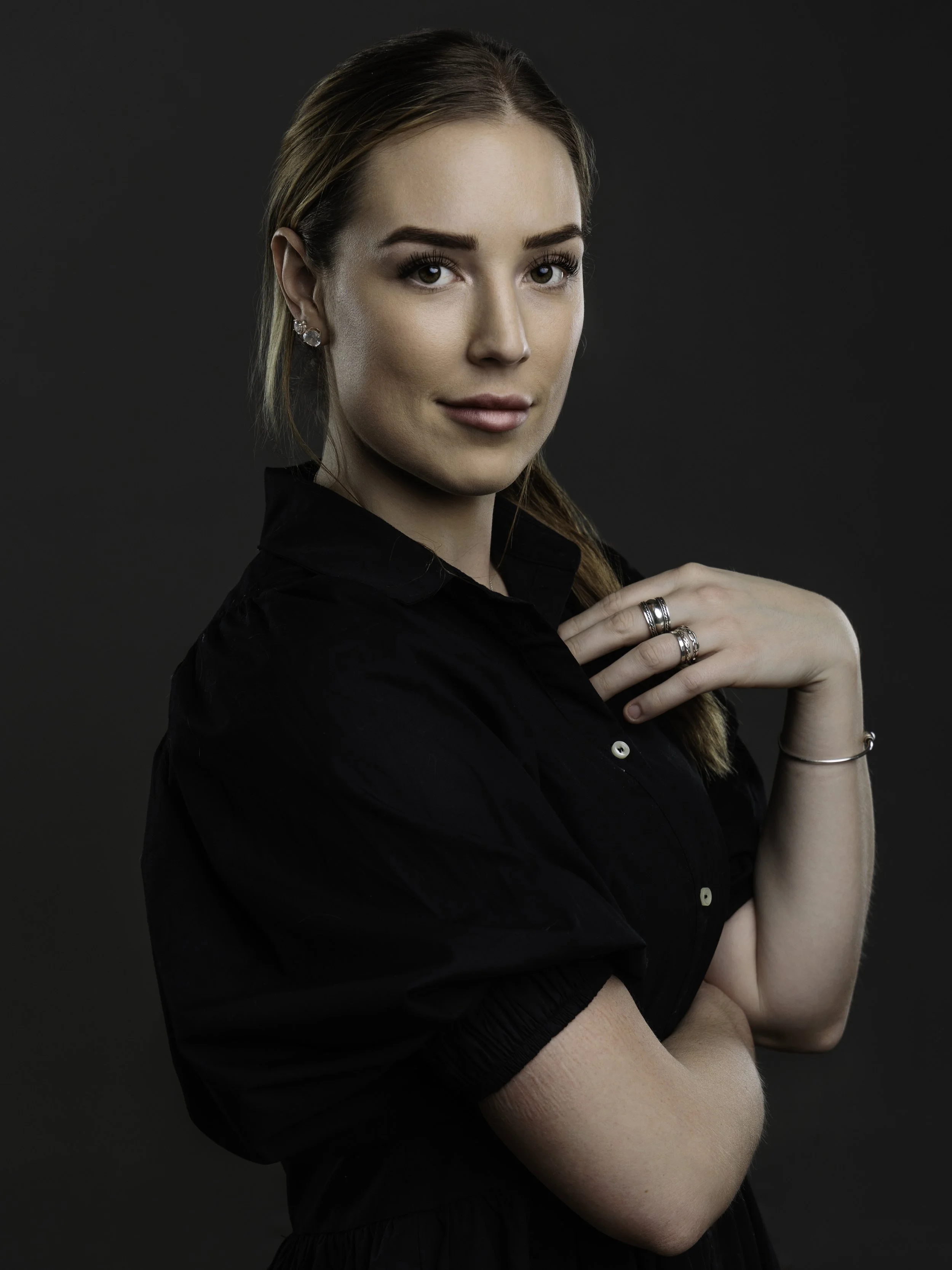 A woman with long brown hair and big brown eyes wearing makeup, jewelry, and a black top posing with her arms crossed against a dark background.