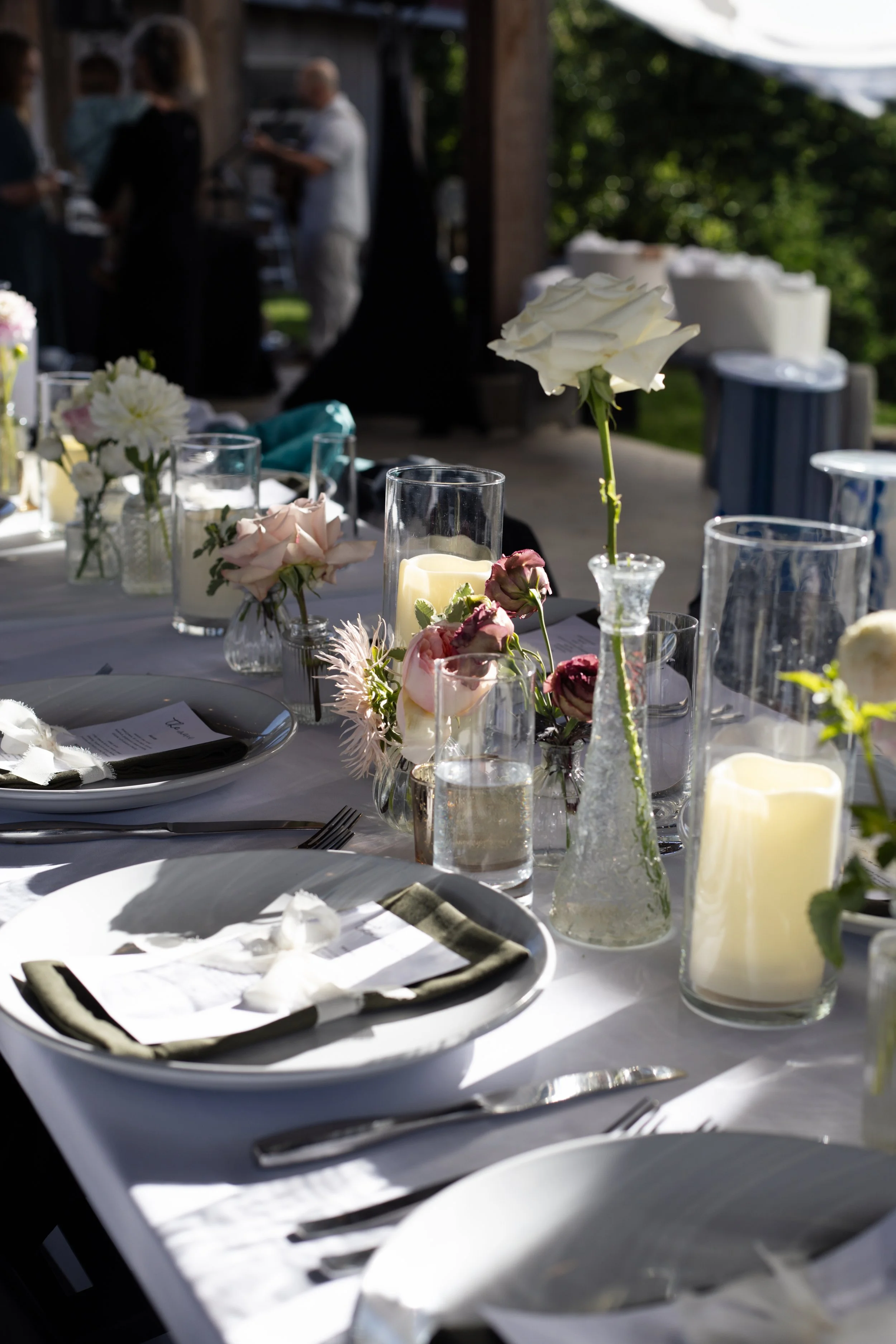 A table set for an outdoor event with floral centerpieces, candles, plates, and silverware. People are in the background, slightly blurred, standing under a picnic shelter on a sunny day.