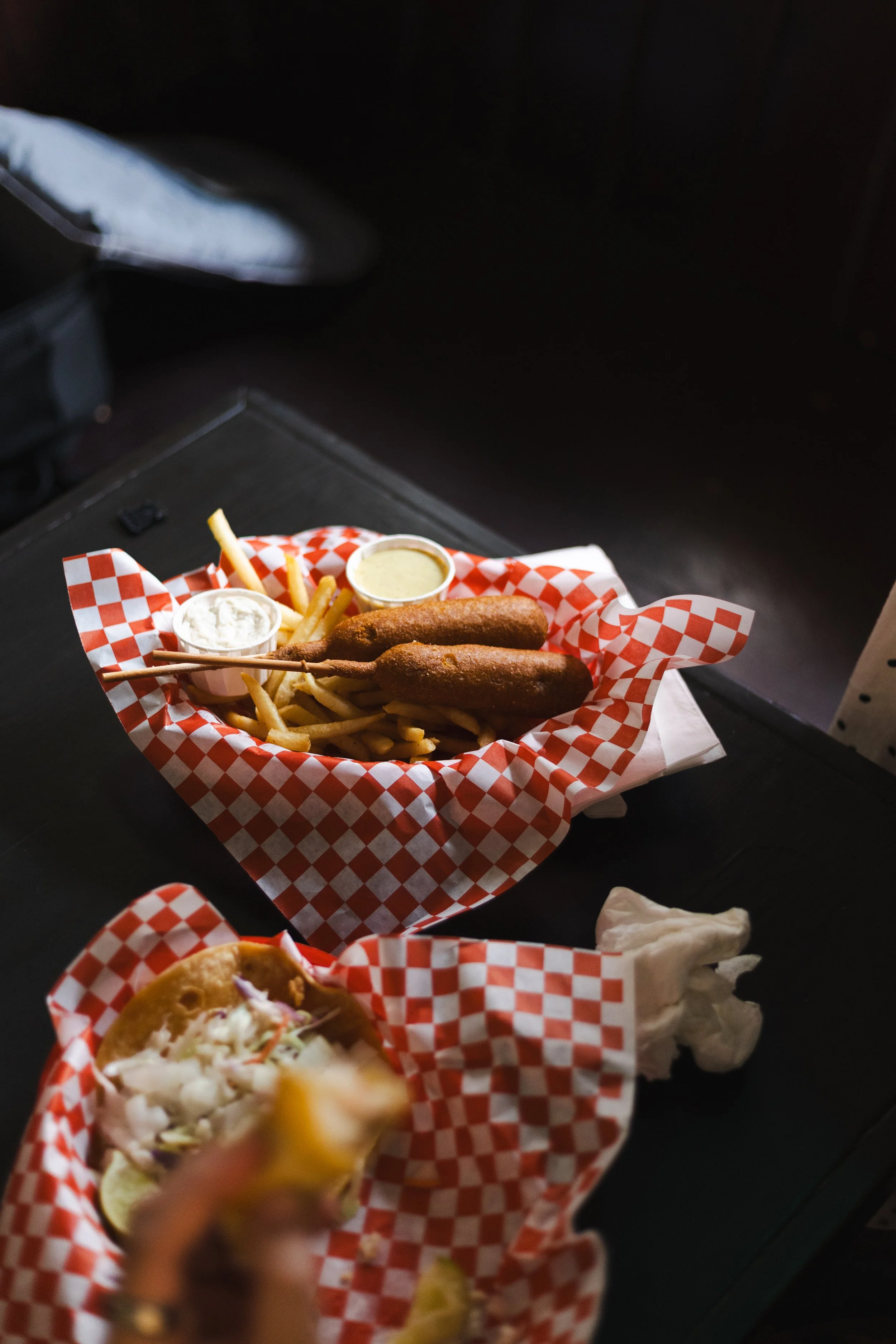 Basket of fried food, including three skewered fried items, French fries, and two dipping sauces on red and white checkered paper.