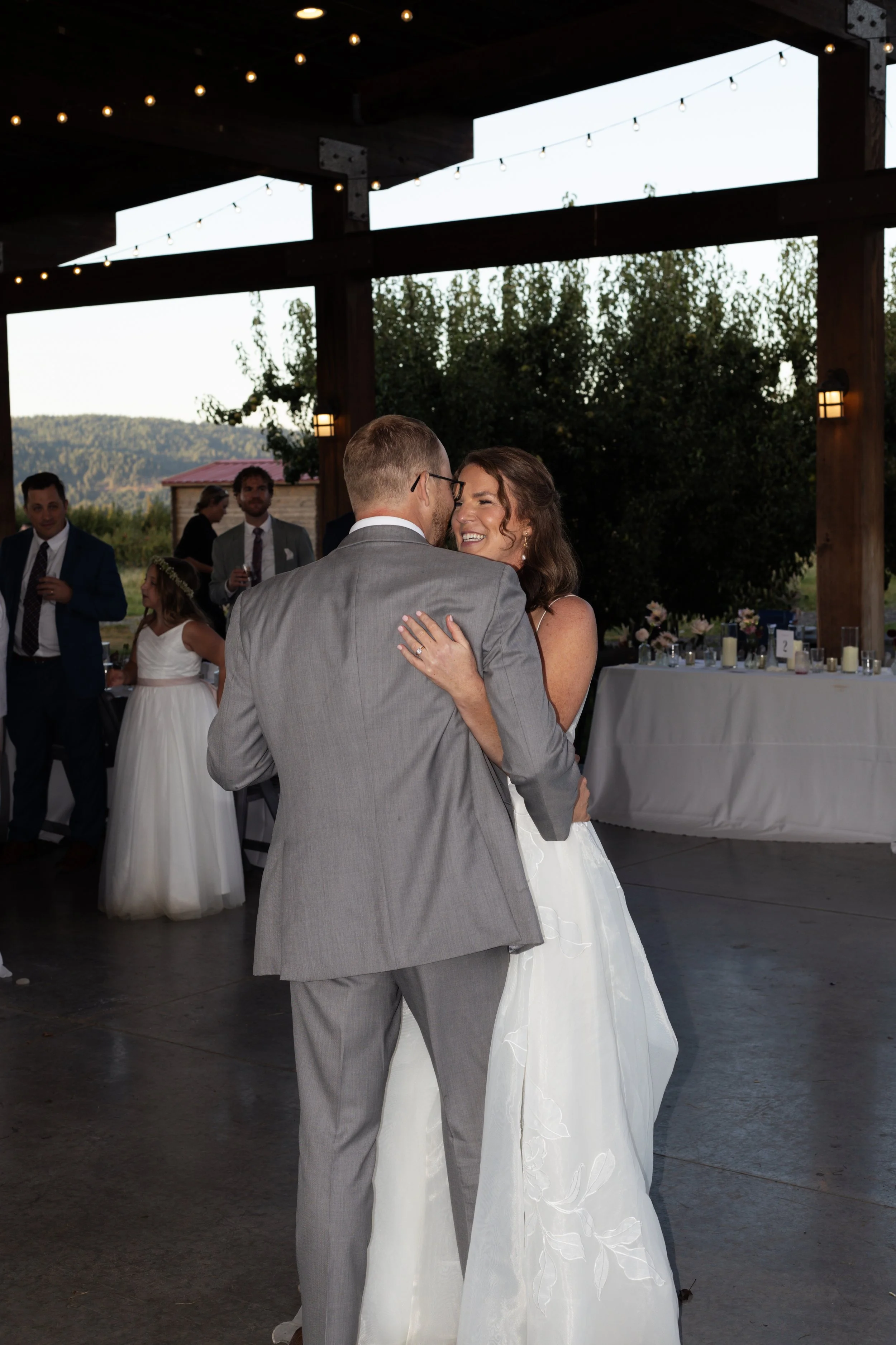 A wedding reception with a bride and groom dancing, surrounded by guests in formal attire, under a wooden pavilion with string lights and outdoor scenery in the background.