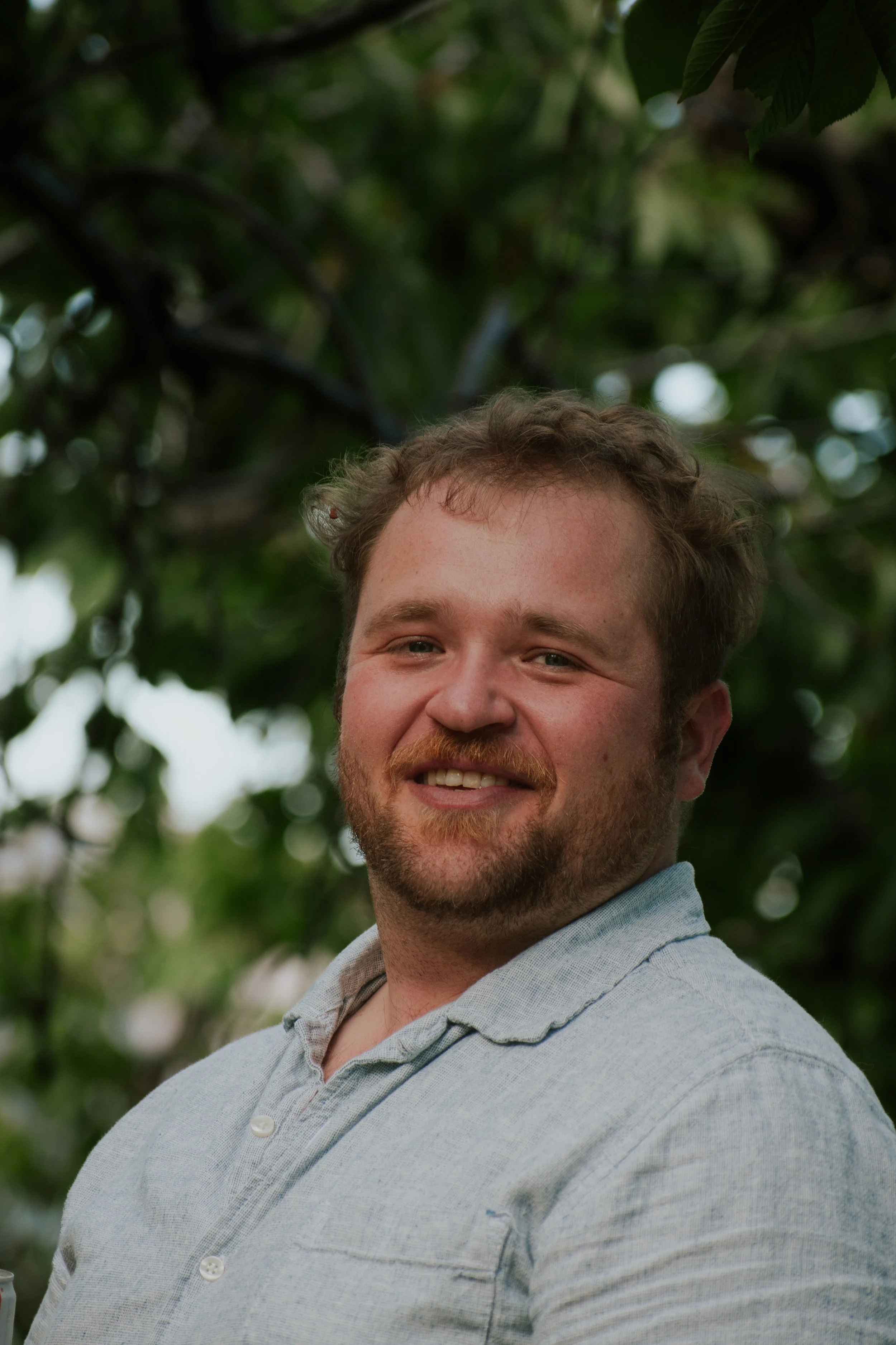 A smiling young man with short curly hair and a beard, wearing a light blue collared shirt, standing outdoors with blurred green foliage in the background.