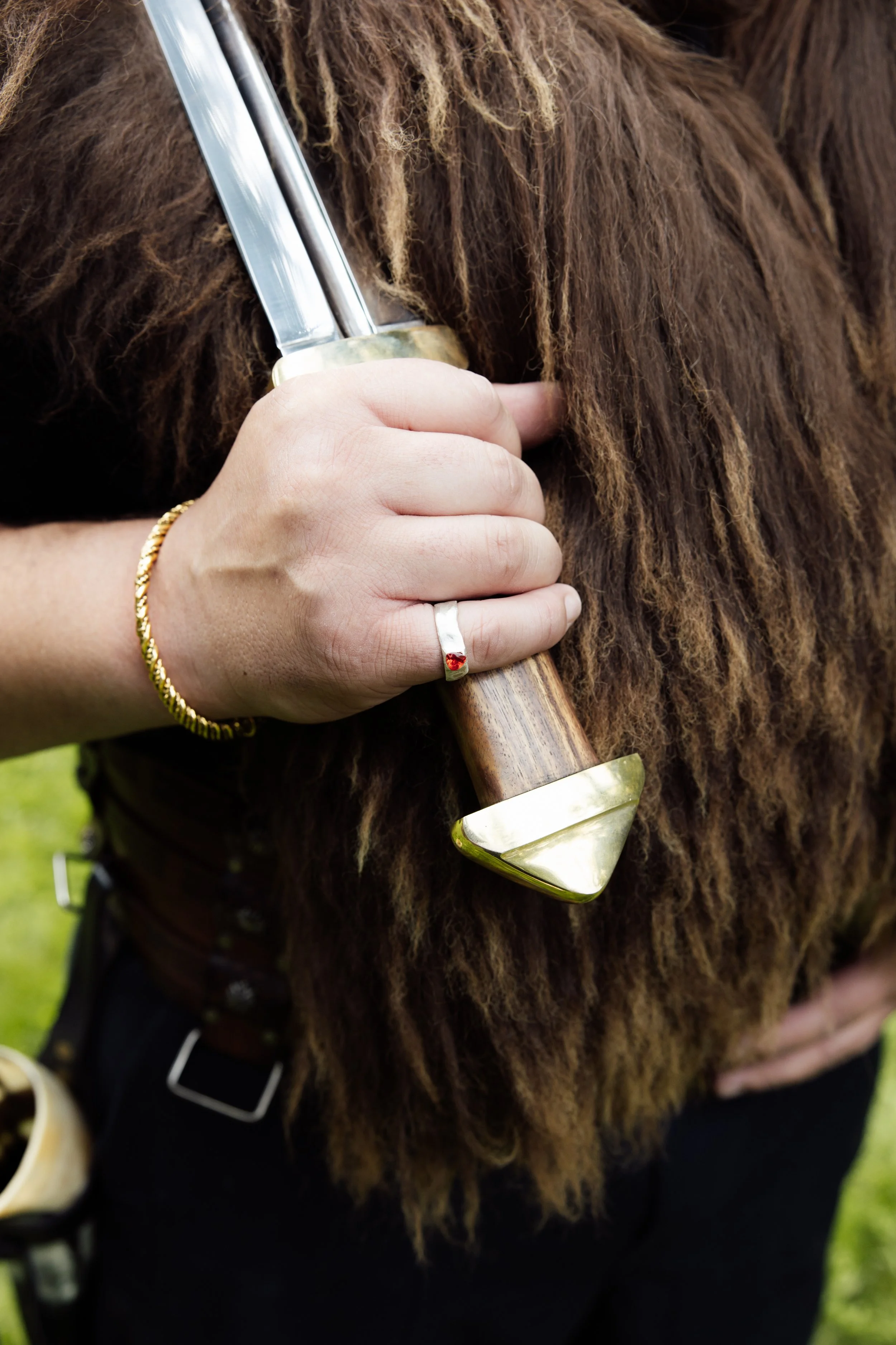 Close-up of a person's hand holding a large knife with a wooden handle, resting on a brown, furry animal hide. The person is wearing a gold bracelet and a silver ring with a small red gemstone.