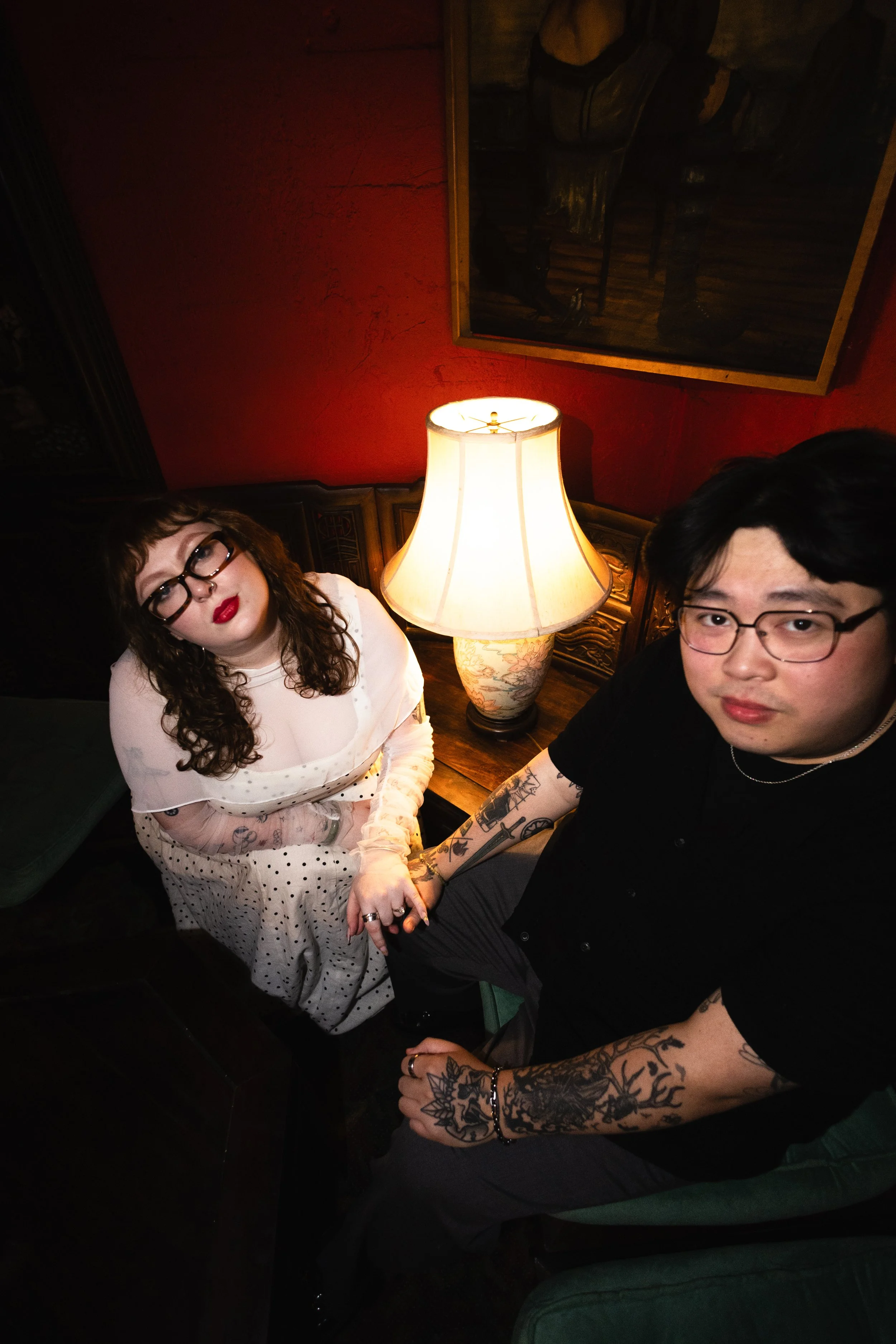 A young woman with curly brown hair, glasses, and red lipstick wearing a white polka dot dress sitting next to a young man with black hair, glasses, and tattoos on his arms, holding hands in a dimly lit room with a lamp and framed paintings on the wa