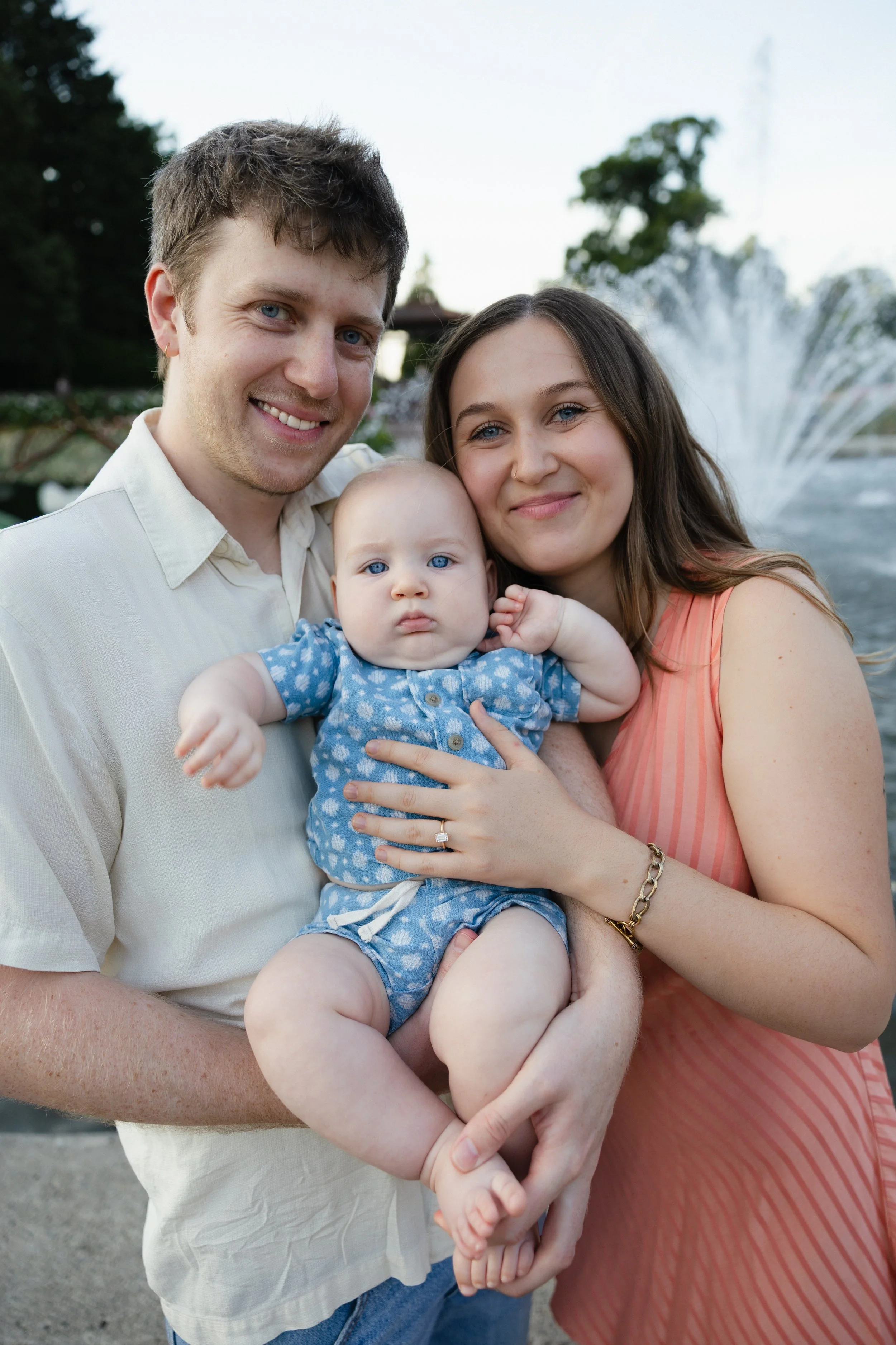A family of three, a man, a woman, and a baby, smiling and embracing outdoors near a fountain with water spray in the background.