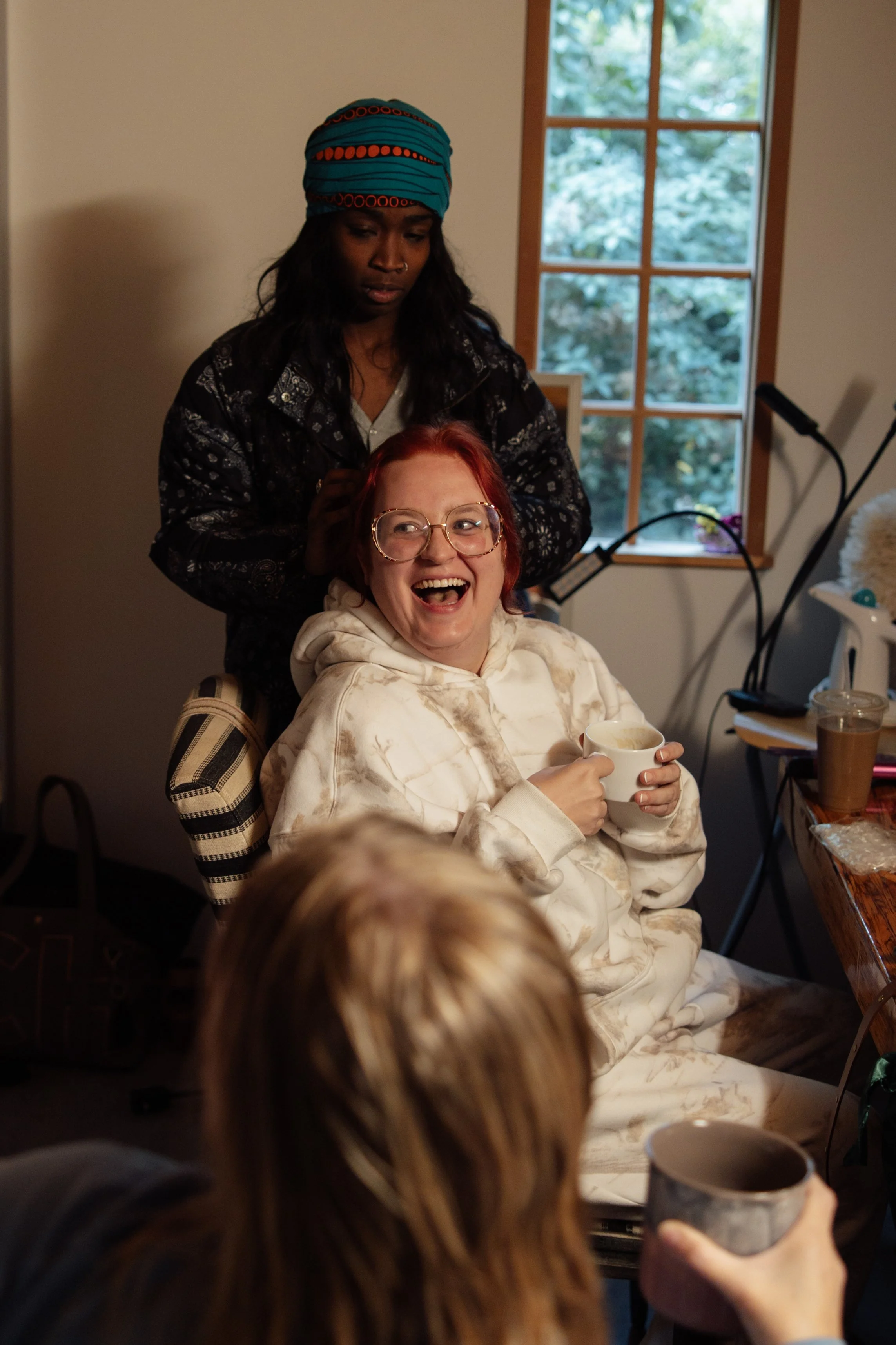 A woman with red hair and glasses sitting in a relaxed pose, smiling and holding a white mug. Behind her, a woman with dark skin wearing a blue headscarf and black patterned jacket stands softly touching her head. The setting appears to be indoors, w