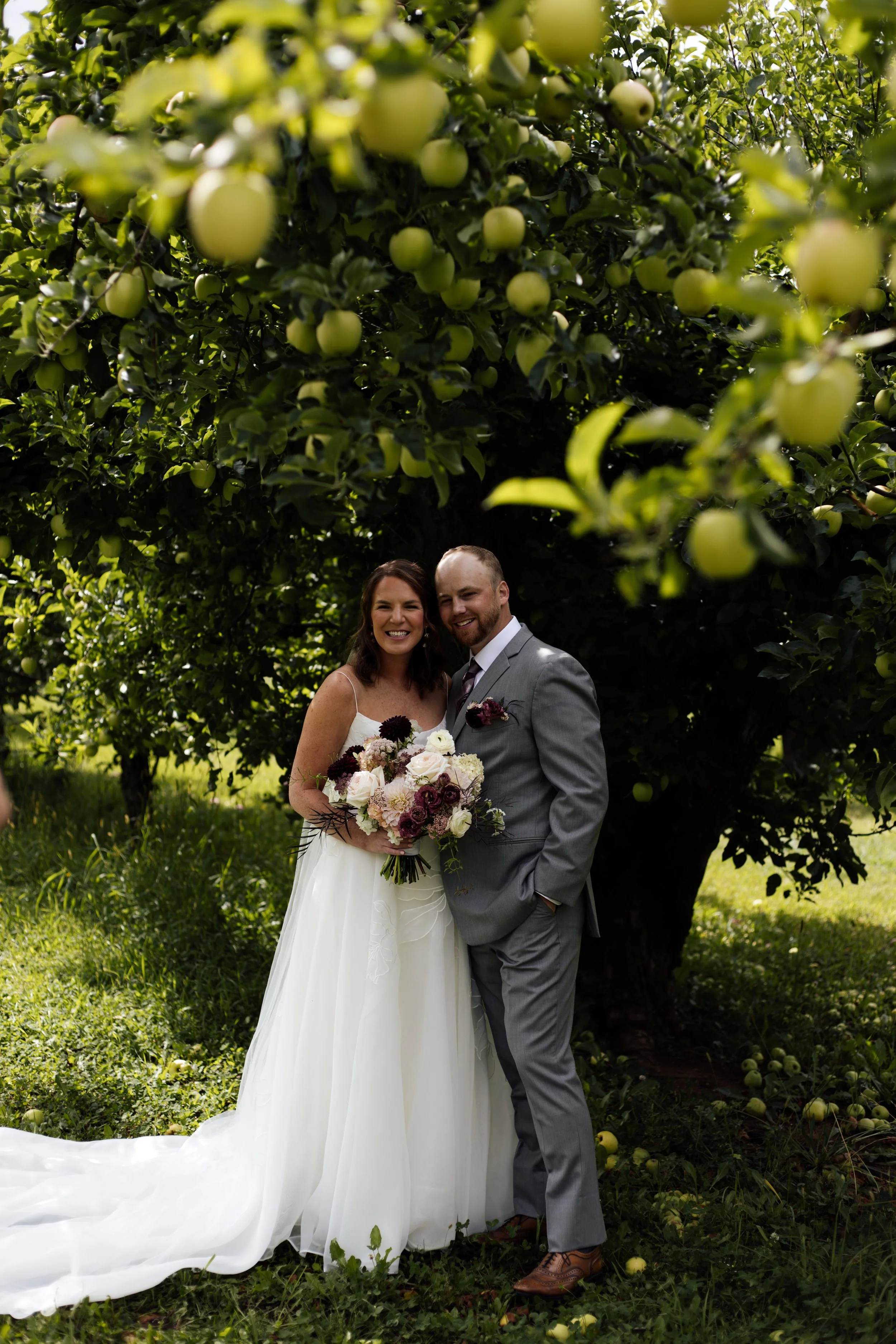 A bride and groom stand together outdoors under a large apple tree with green apples, smiling at the camera. The bride wears a white wedding dress and holds a bouquet of flowers. The groom wears a gray suit with a tie and brown shoes.