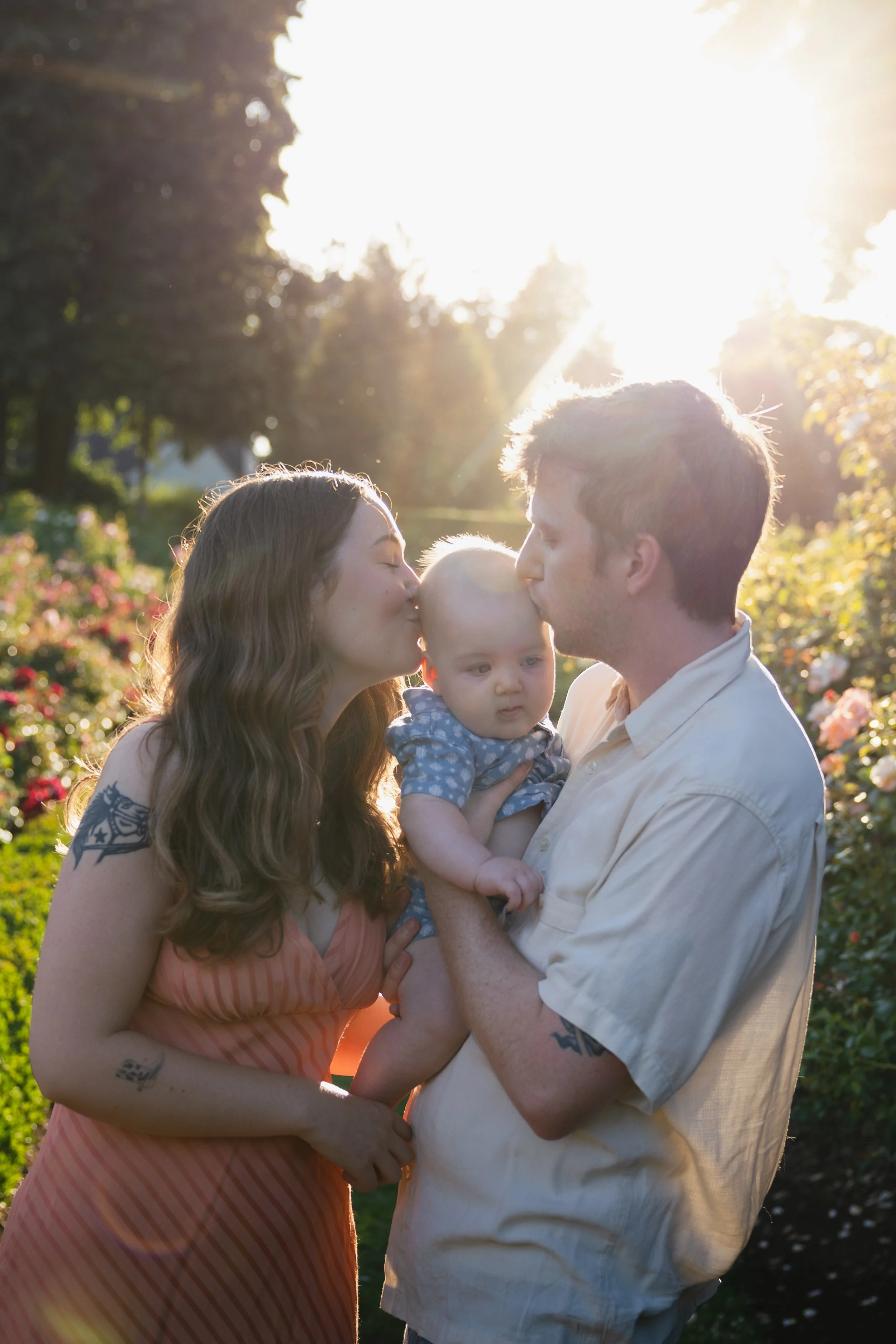 A young woman and a young man are kissing a baby on the cheeks outdoors in a garden with trees and flowers, illuminated by bright sunlight.