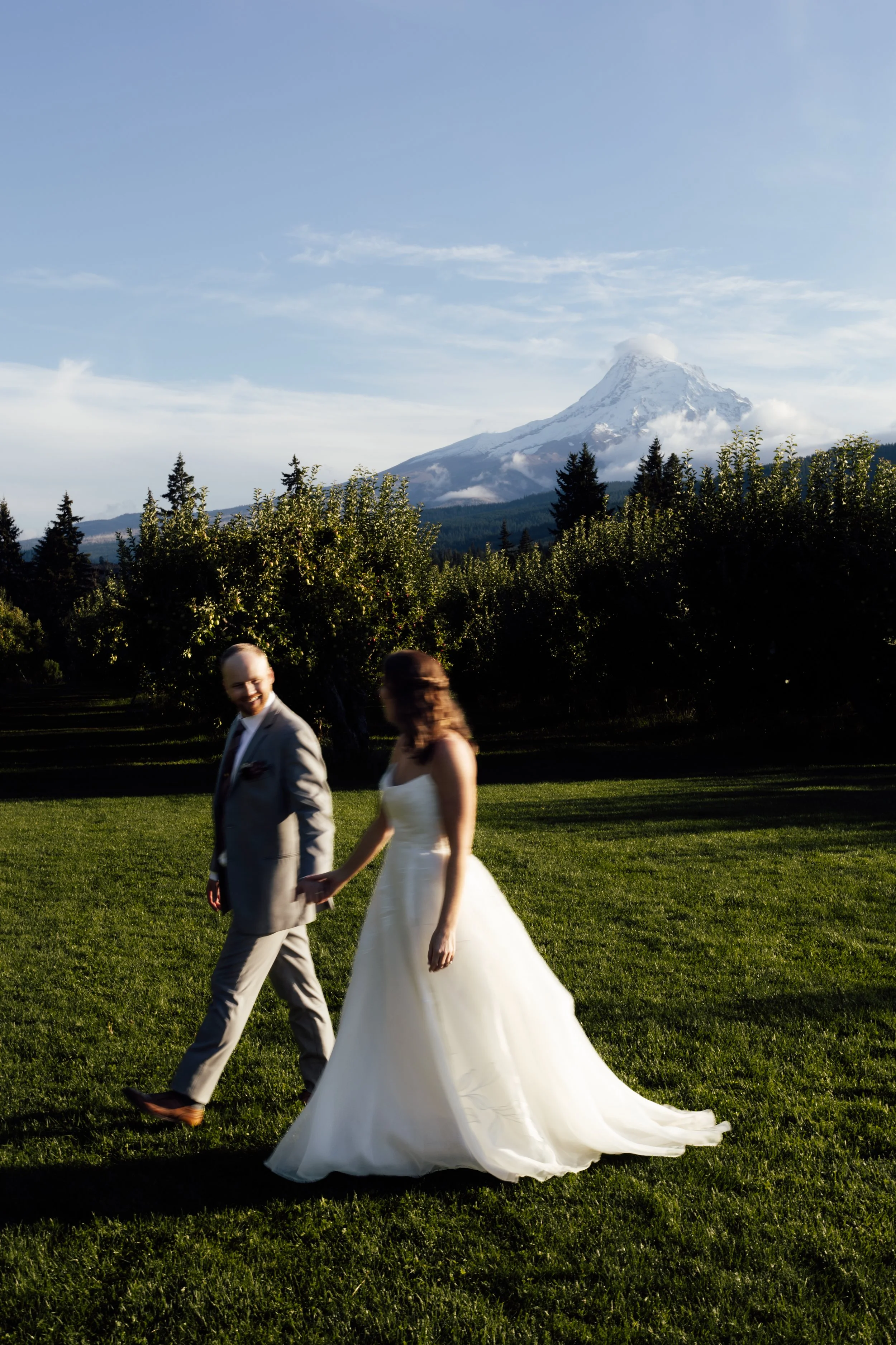 A bride and groom walking hand in hand on a grassy field with Mount Hood, snow-capped, in the background during daytime.