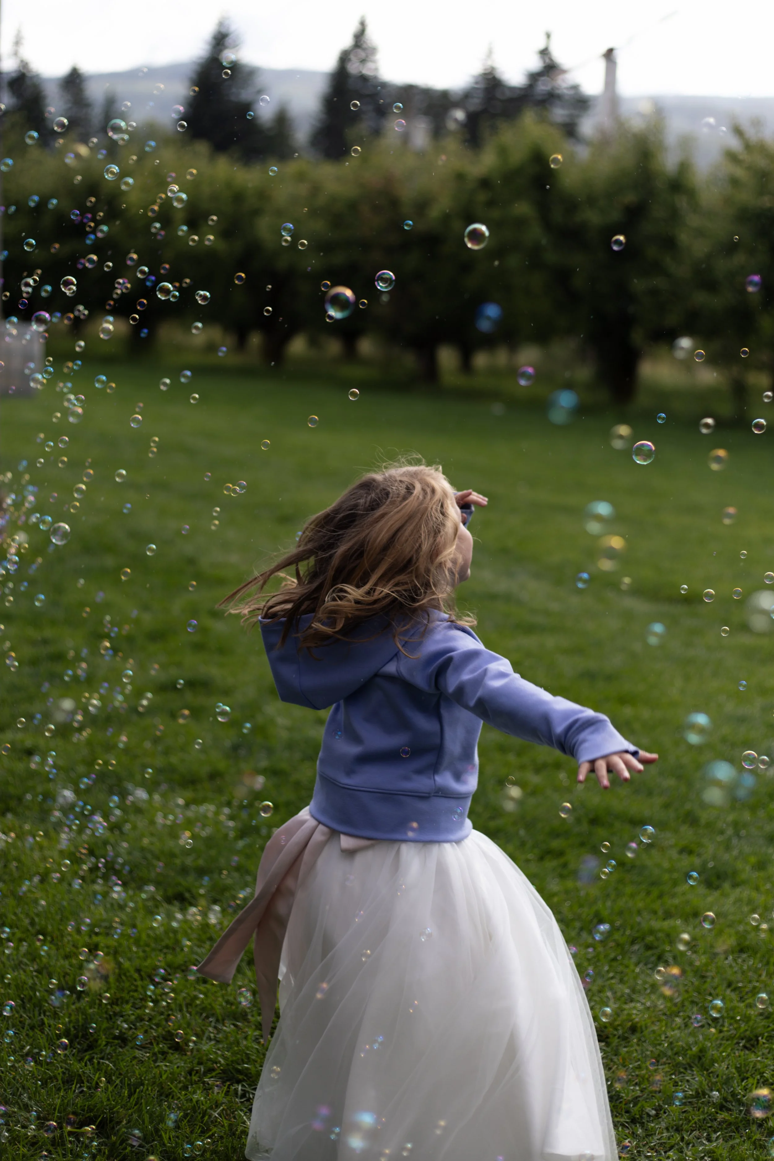 Girl in a blue hoodie and white tulle skirt playing with soap bubbles in a grassy park, with trees and hills in the background.