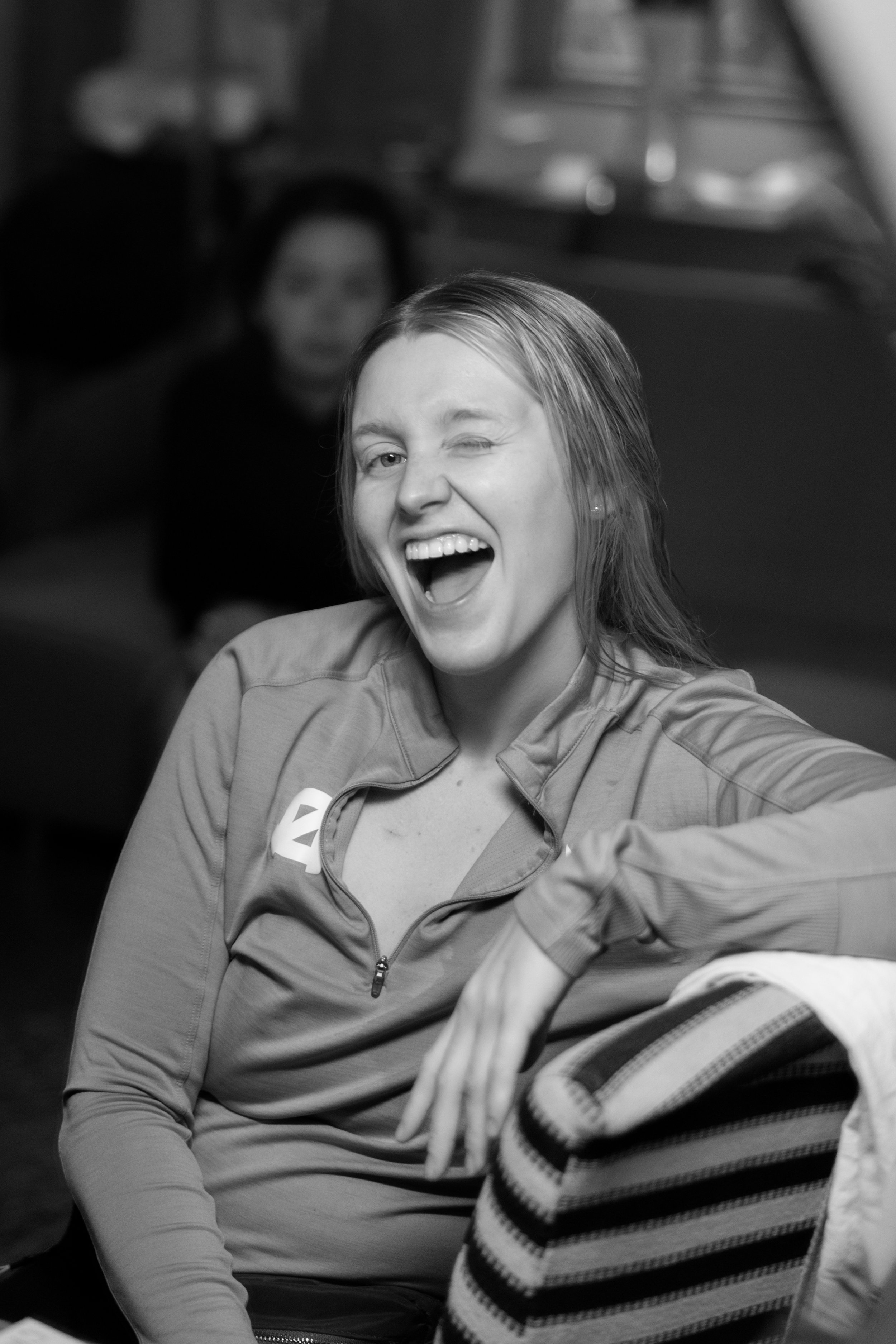A young woman with light hair is smiling and winking at the camera. She is wearing a zip-up jacket and sitting in a casual setting.