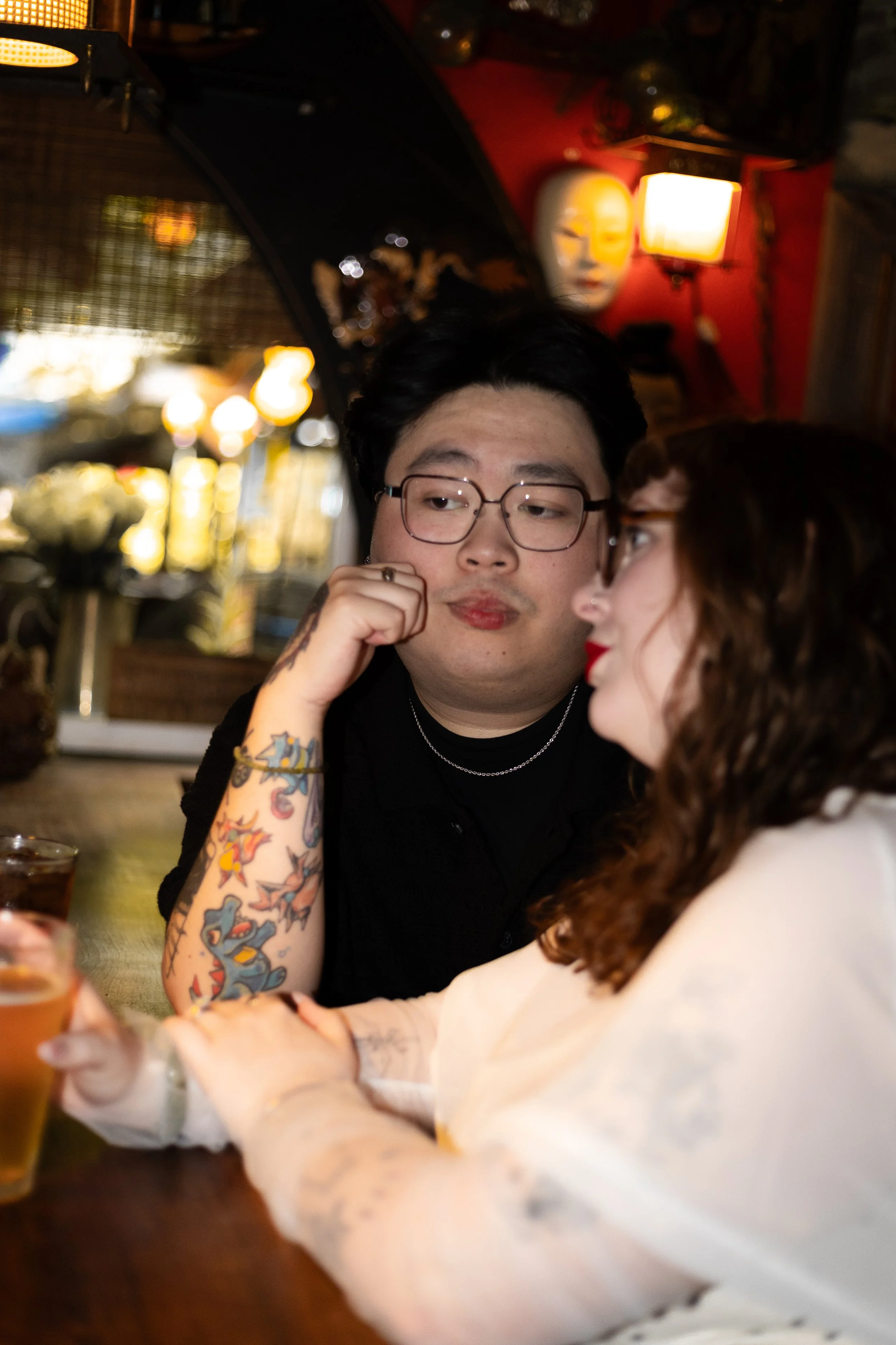 A man wearing glasses with tattoos on his arm sitting at a table, engaging in conversation with a woman who has fair skin and long curly hair. The background features dim lighting and Asian decorative elements.