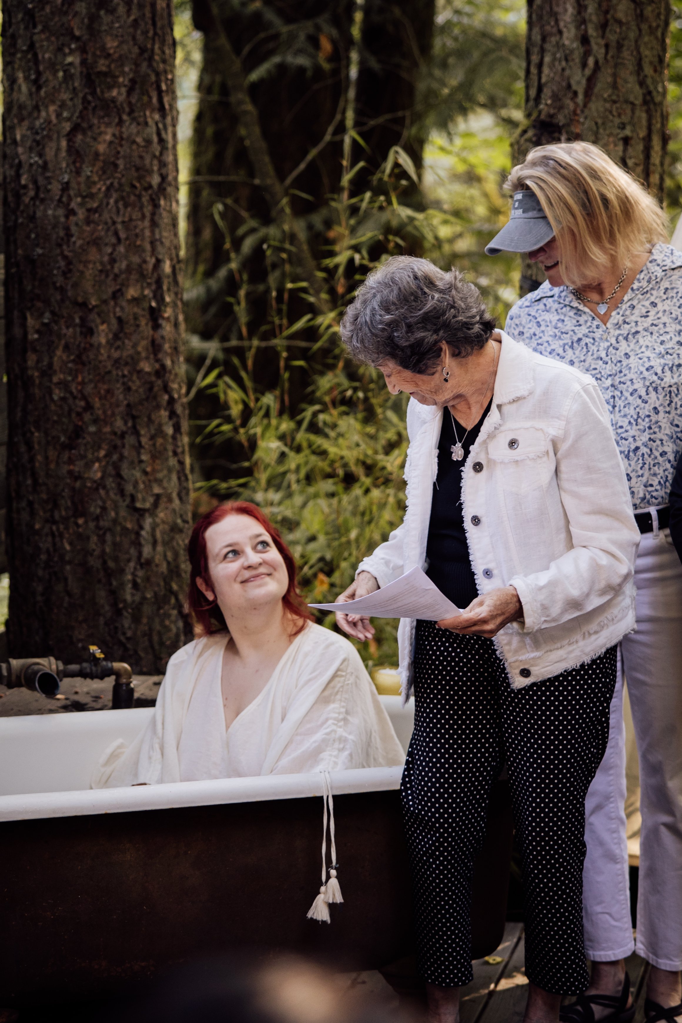 A woman with gray hair and a white jacket talking to a younger woman with red hair seated in a bathtub outdoors in a forest setting.