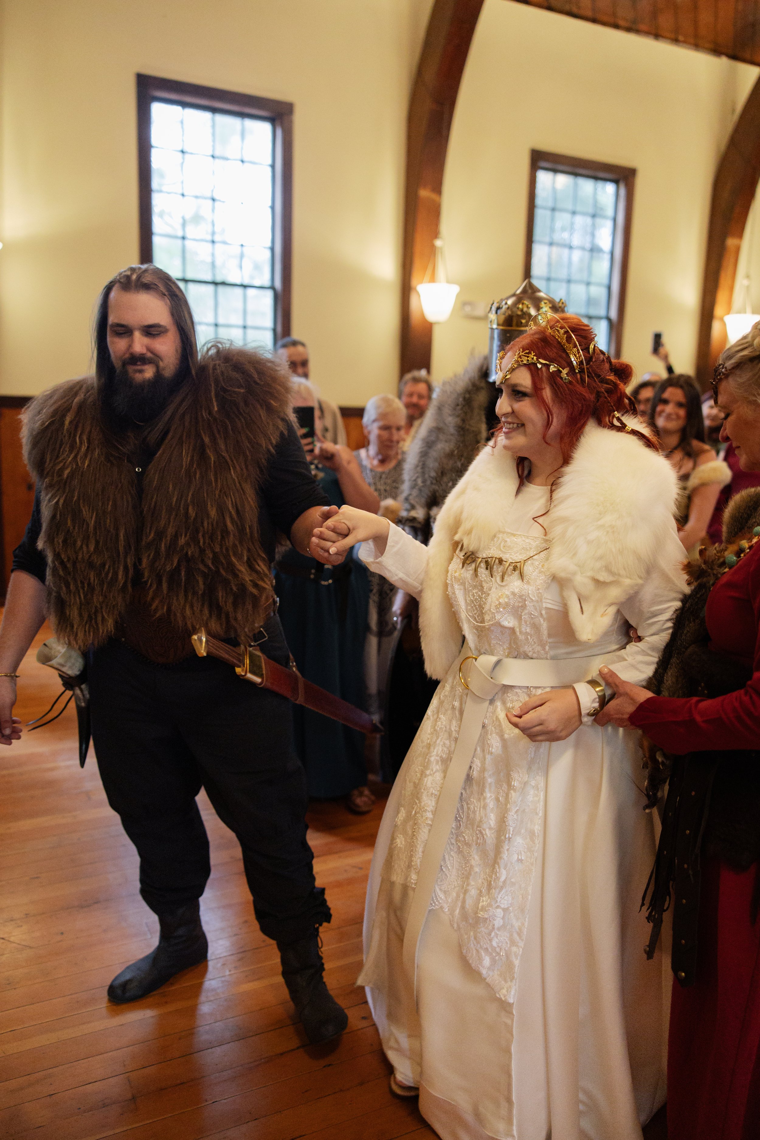 A couple dressed in medieval or fantasy costumes, dancing at an indoor event with wooden floors and large windows, surrounded by people watching and taking photos.