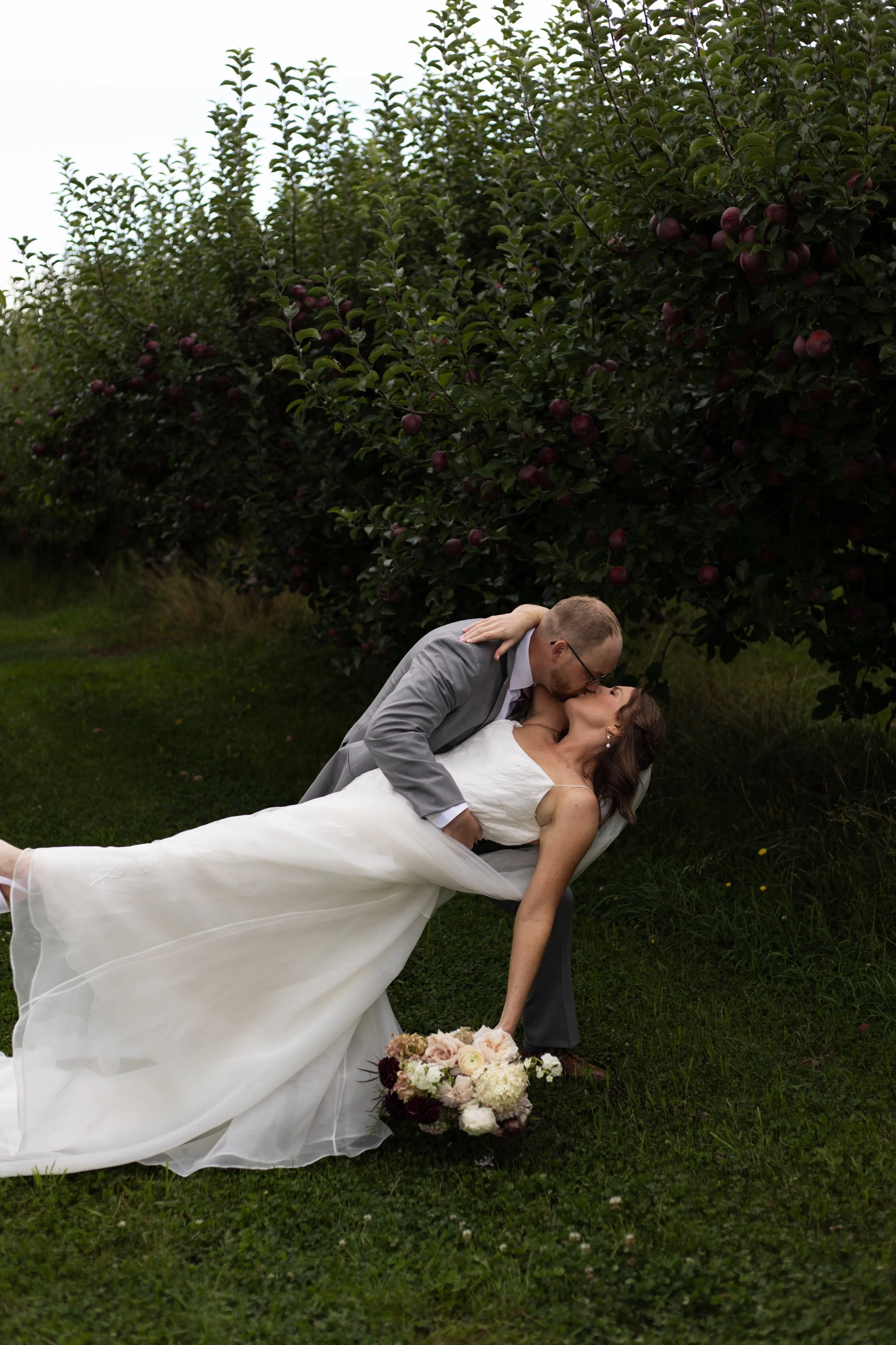 A newlywed couple sharing a kiss outdoors during their wedding, with the groom bending down and the bride lying on the grass, holding a bouquet of flowers, against a backdrop of lush green trees and apples.