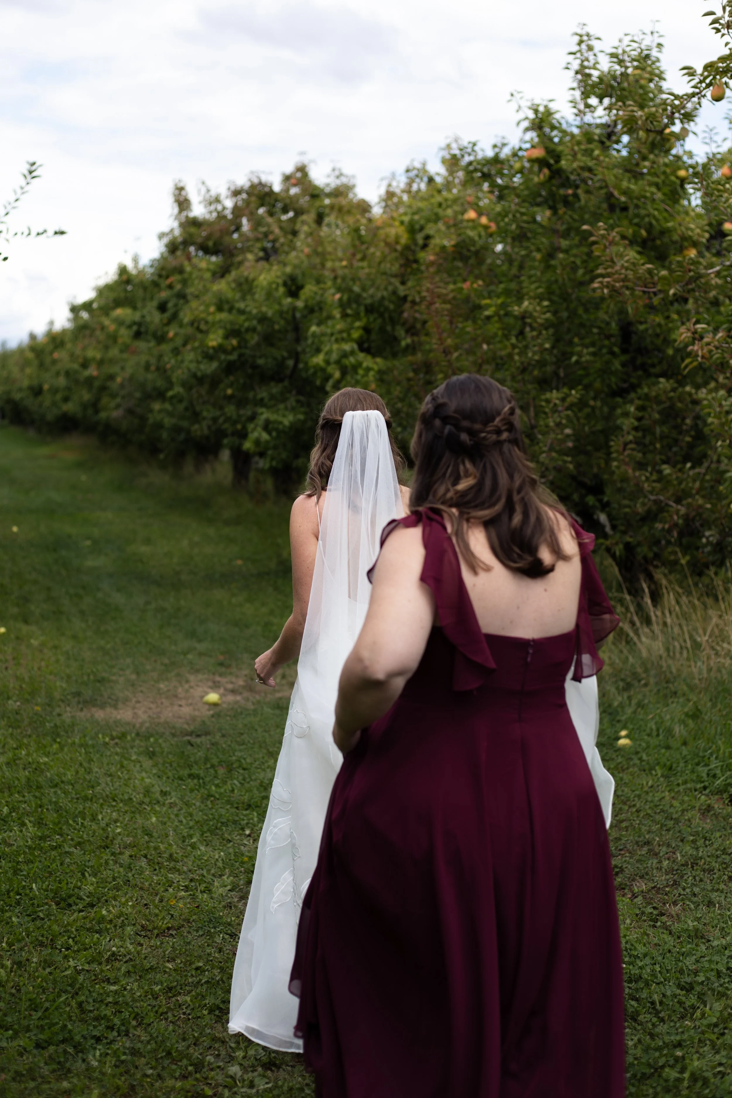 Two women walking in a garden with trees, one in a white wedding dress with a veil, the other in a maroon dress with braided hair.