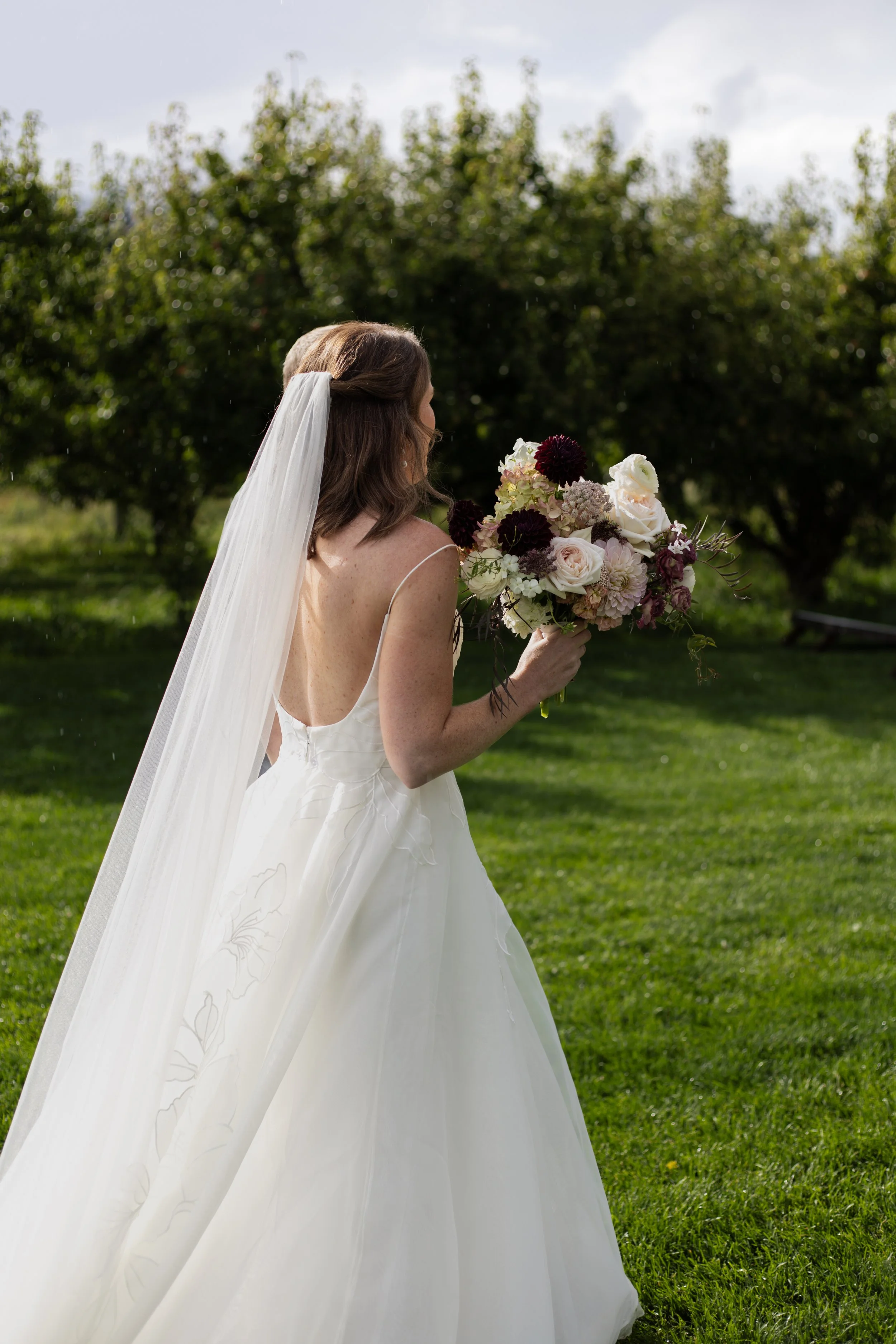 Bride in a white wedding gown with spaghetti straps and a long veil holding a large bouquet of white and dark purple flowers outdoors.