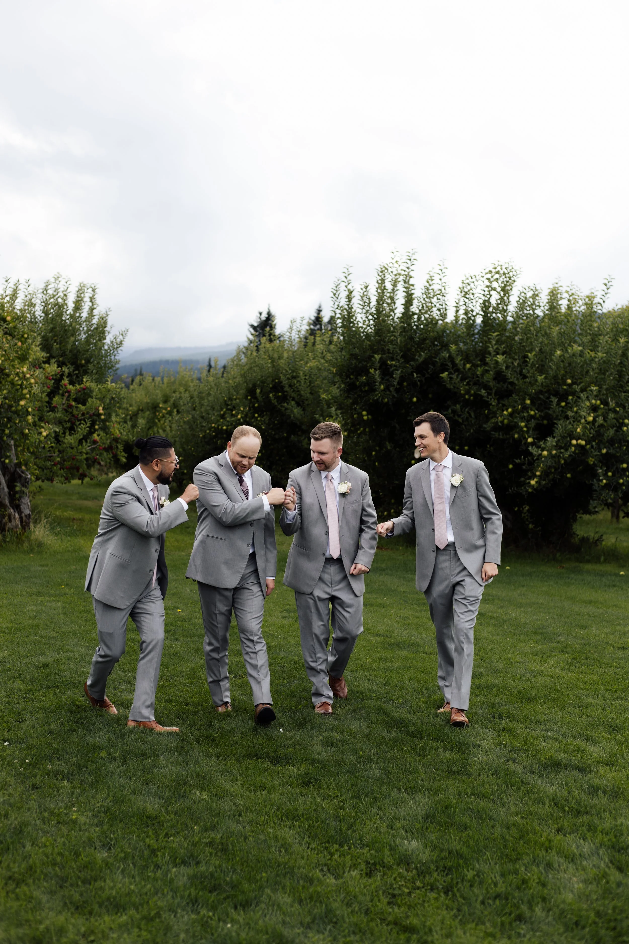Five men in gray suits holding hands and walking together on a grassy field outdoors, smiling and engaged in conversation, with trees and hills in the background under a cloudy sky.