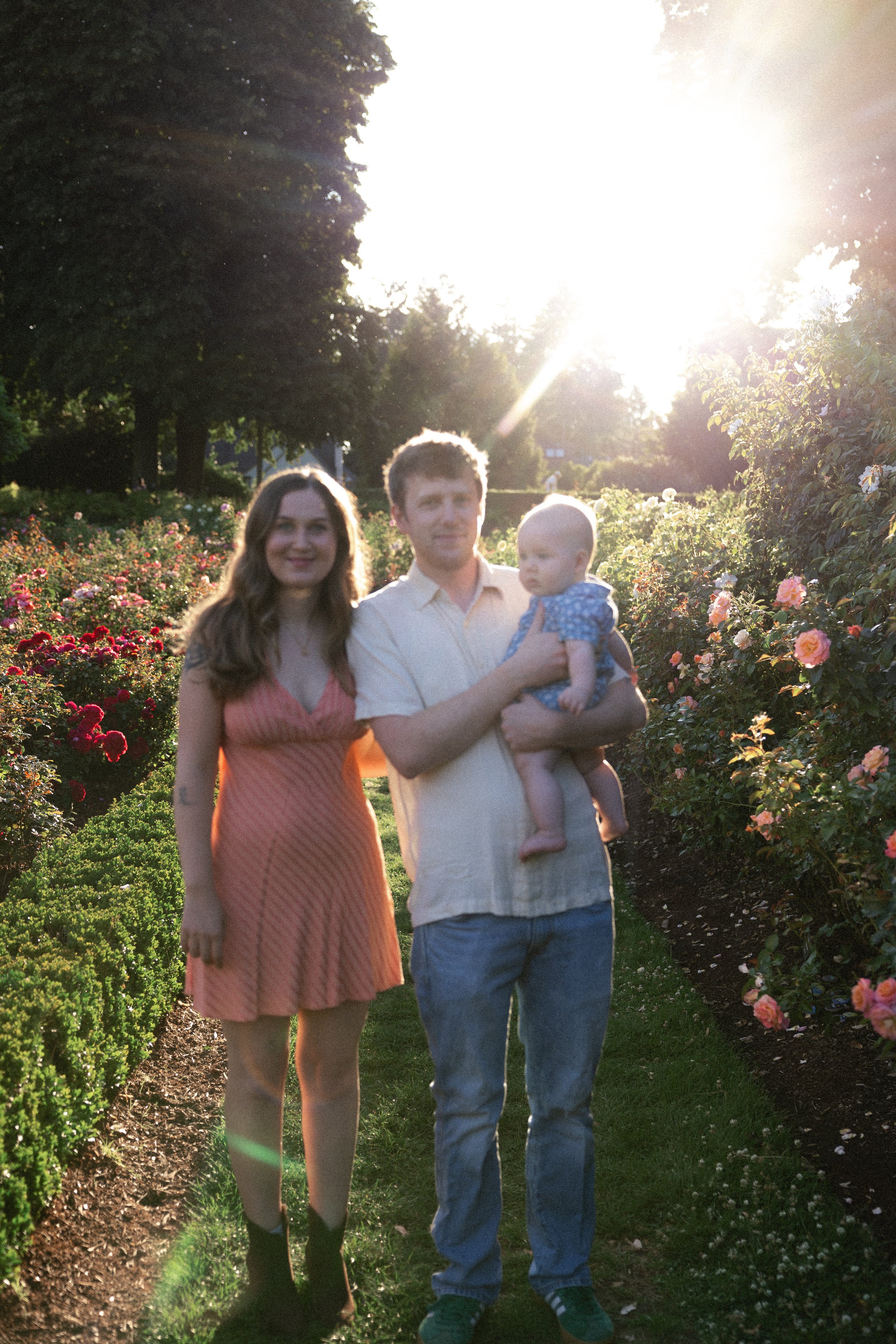 A family of three posing outdoors in a garden with blooming roses during sunset. The woman is wearing a pink dress with black boots, the man is holding a baby and wearing a light-colored shirt and blue pants, and they are standing among rose bushes w