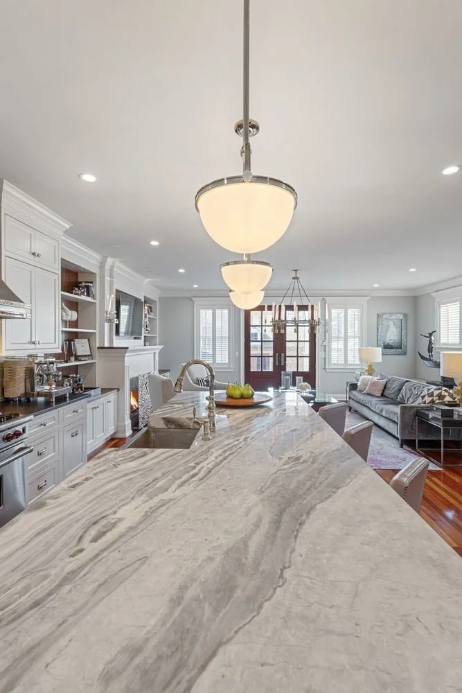 Modern open-concept kitchen and living room with a marble island, pendant lighting, white cabinetry, and large windows.