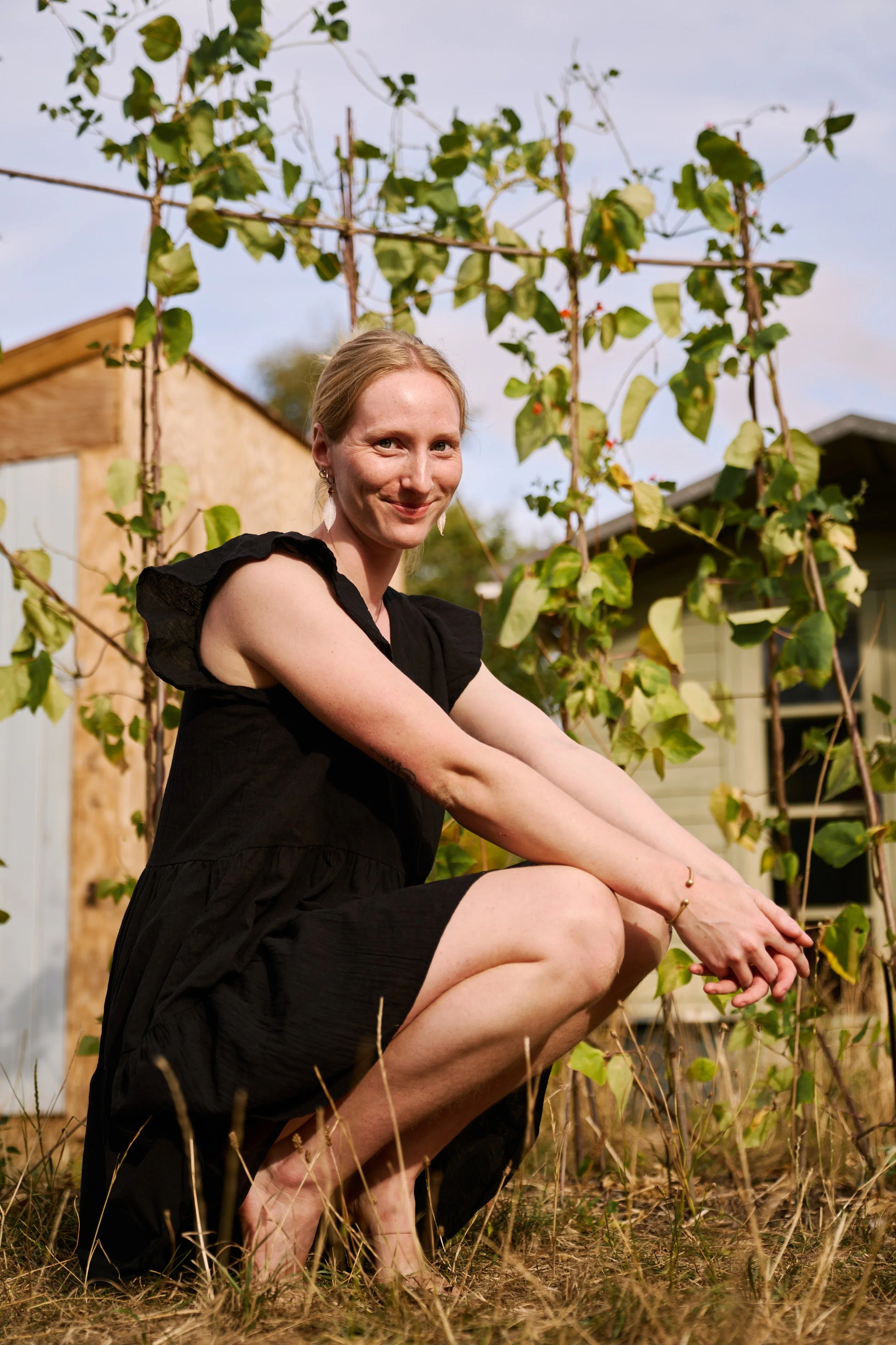 Woman with blonde hair and earrings, wearing a black dress, crouching outdoors in a garden with dried grass and a vine trellis behind her, smiling at the camera.