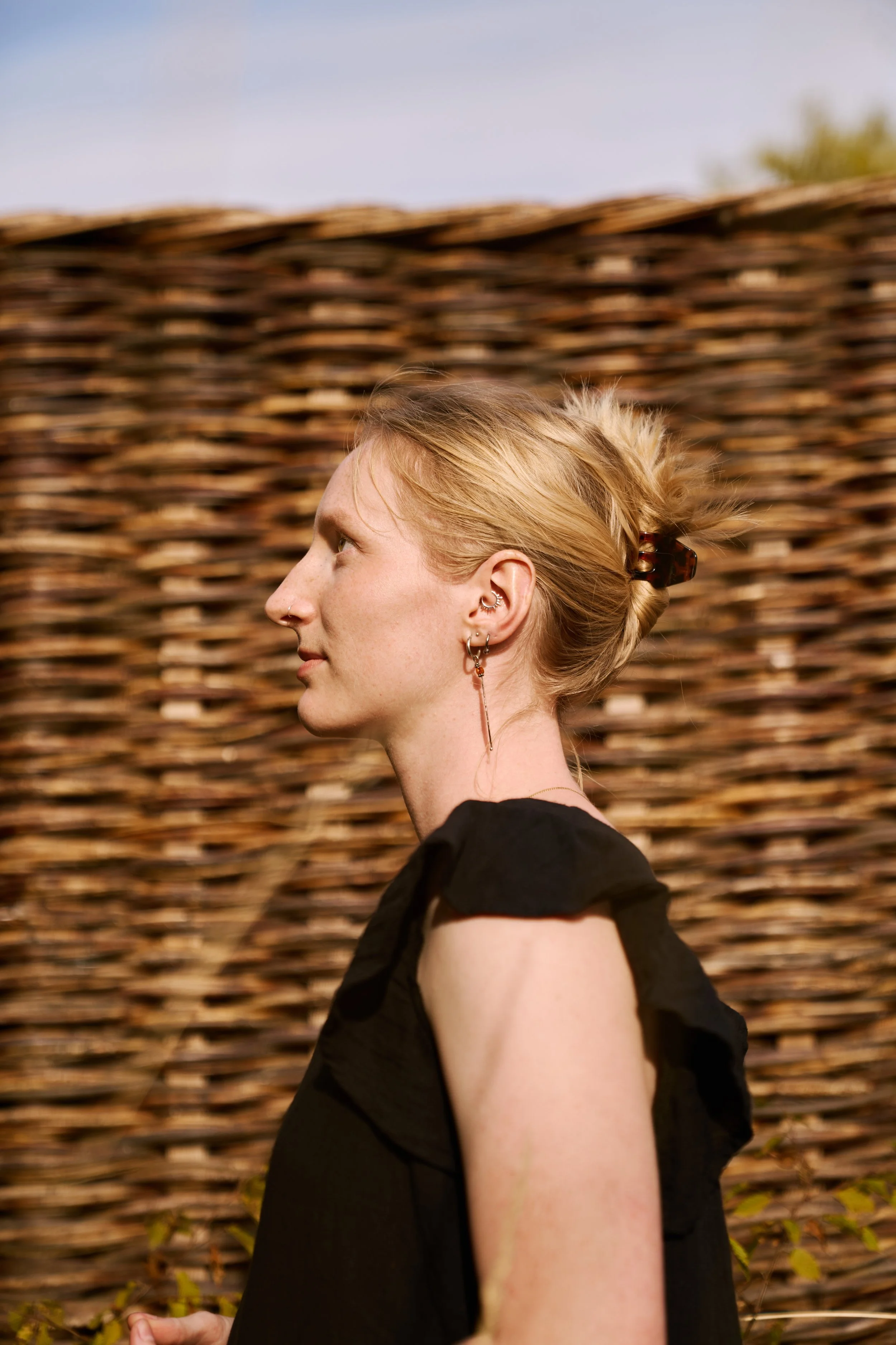Profile of a woman with blonde hair tied in a claw clip, standing against a woven willow fence outdoors.