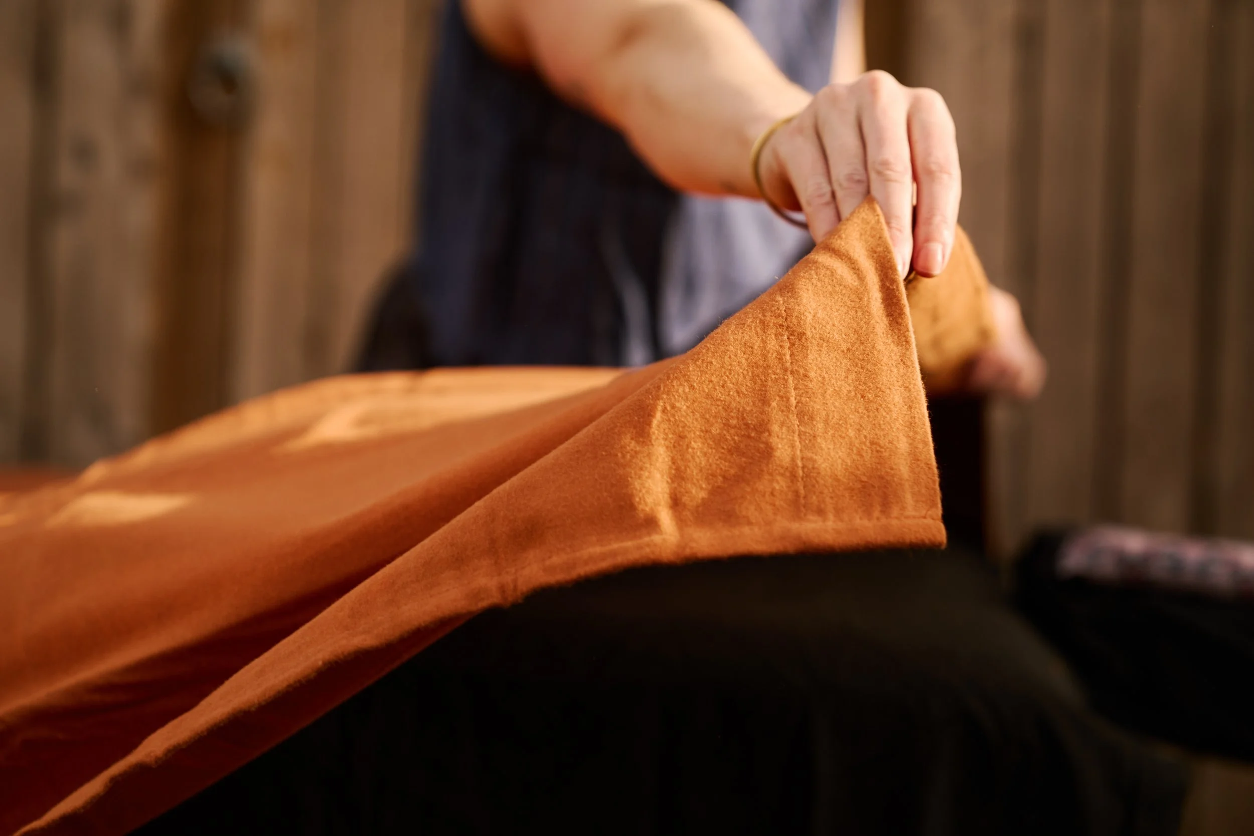 A person holding a folded orange fabric drape for massage with a wooden wall in the background.