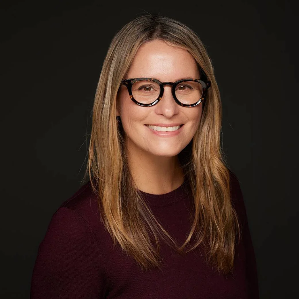Jen Wallace in tortoiseshell glasses smiling against a dark background