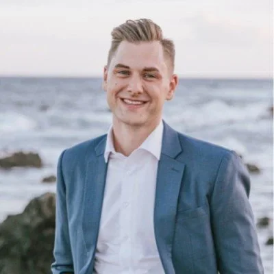 Daniel Hettwer in a blue suit smiling on a beach with rocks and ocean behind him
