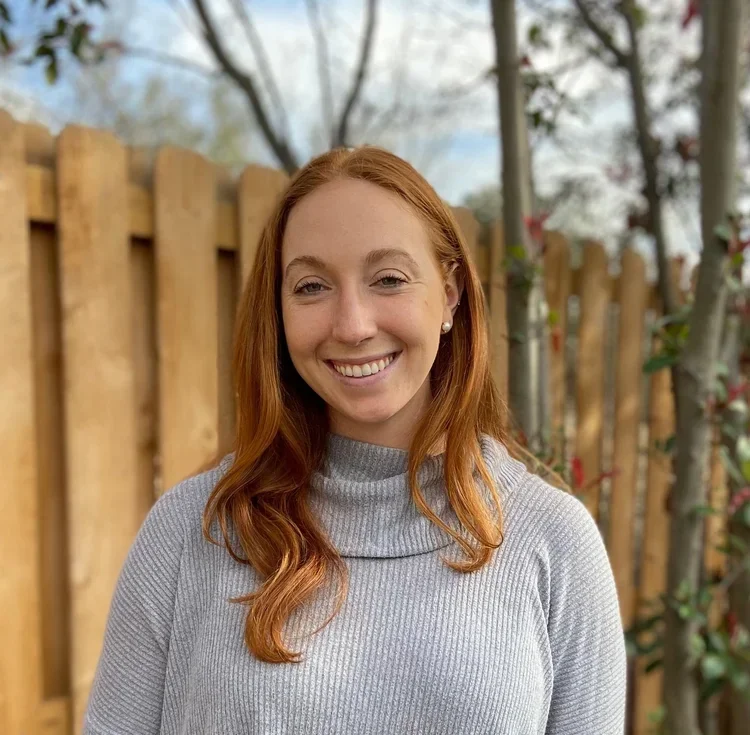 Amy Wentzel smiling outdoors in front of a wooden fence and trees
