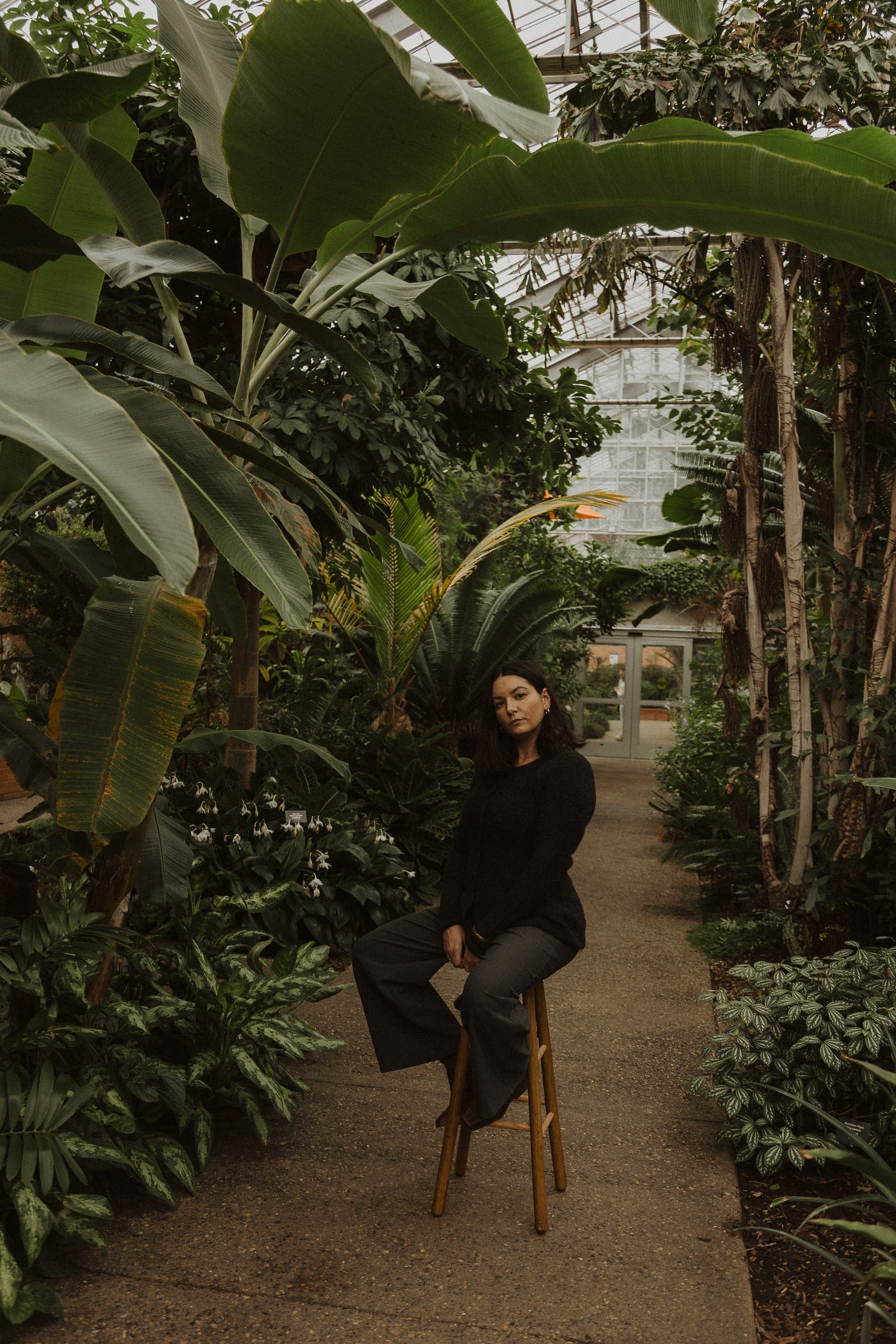 A woman with dark hair seated on a stool in a lush, green greenhouse filled with various tropical plants and trees, with some flowers visible among the foliage.