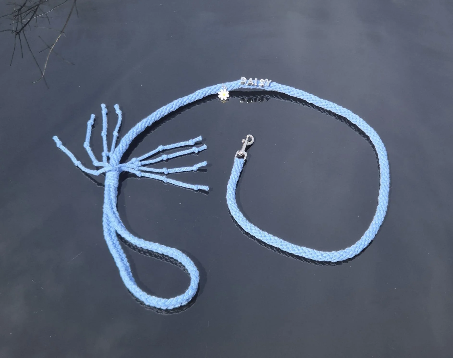 A blue braided necklace with silver charms, one spelling 'DAISY' and a small flower charm, laid on a dark surface.