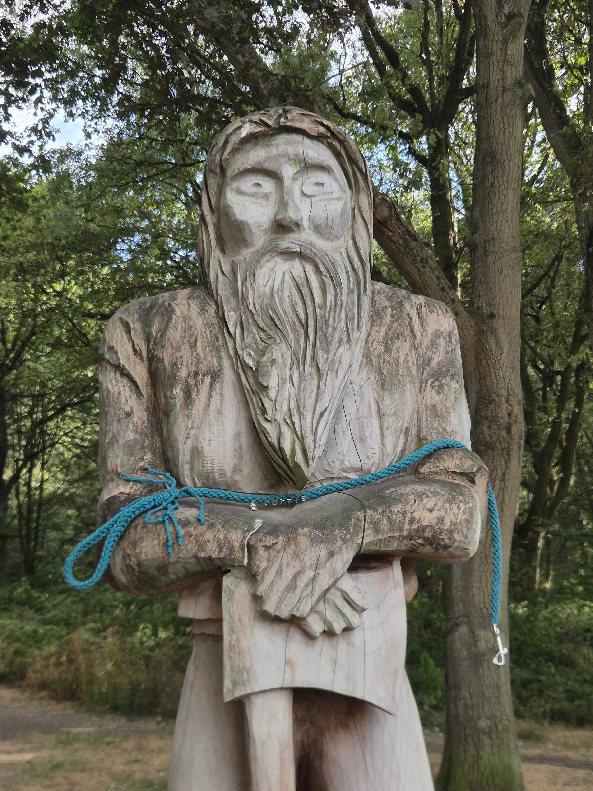 Wooden statue of a bearded man with long hair, sitting outdoors among trees, with a dog lead around its shoulders.