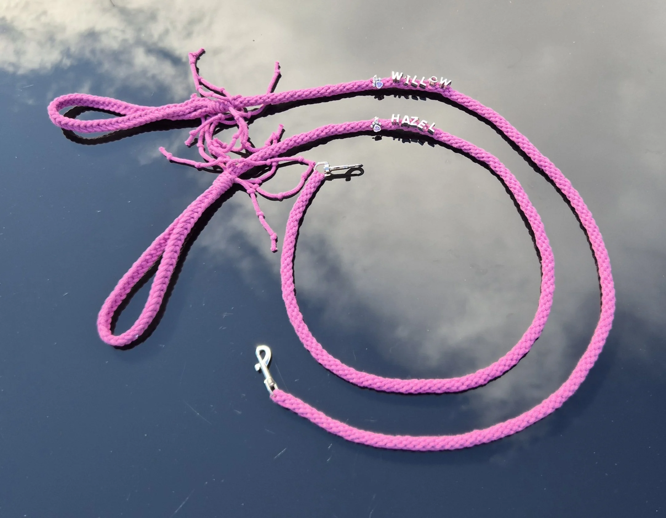 Pink braided necklace with silver charms spelling 'Willow' and 'Hazel' on a reflective surface.