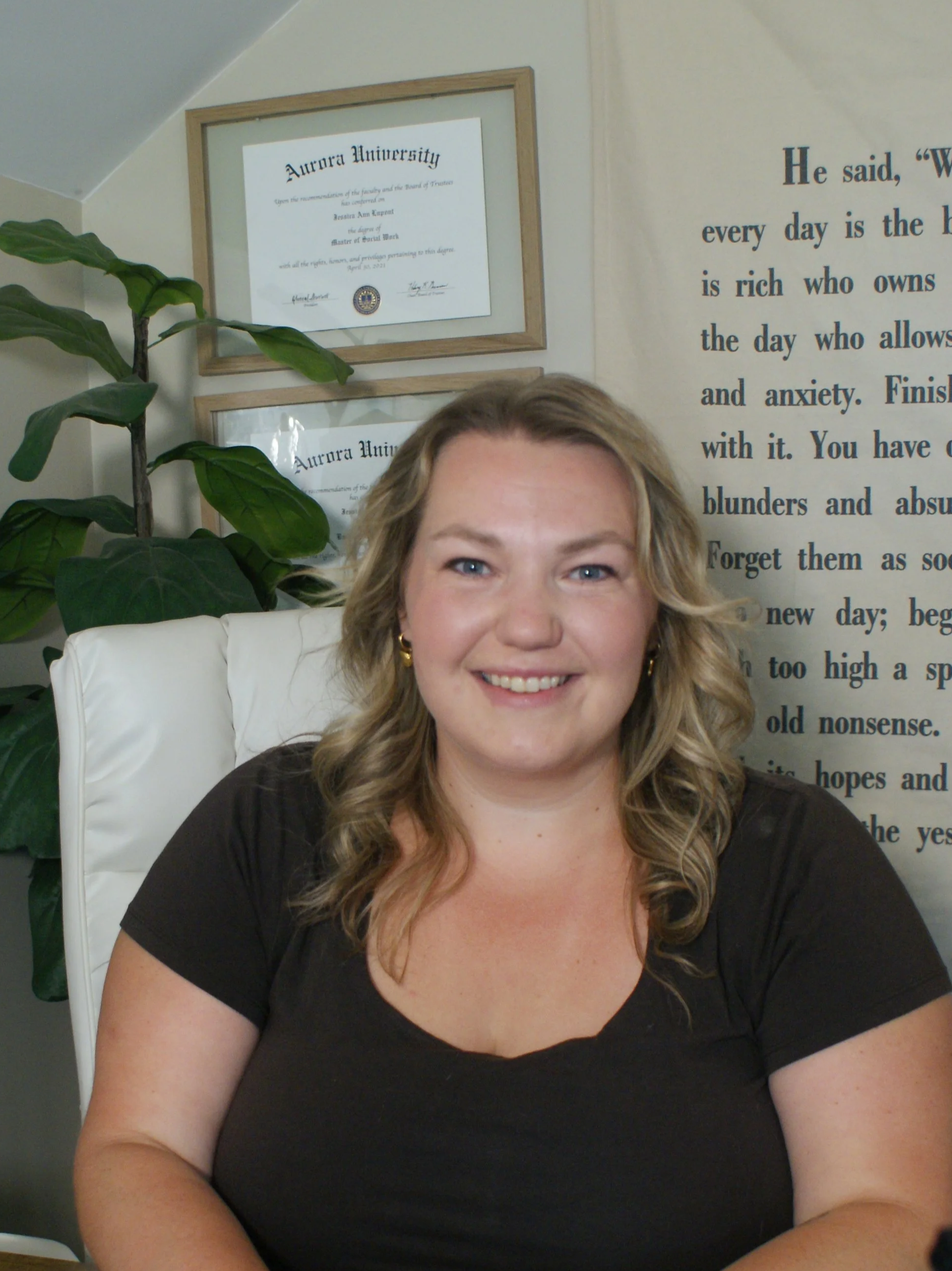 A smiling woman with blonde, wavy hair sitting in a white chair in an office setting, with framed diplomas from Aurora University on the wall behind her.