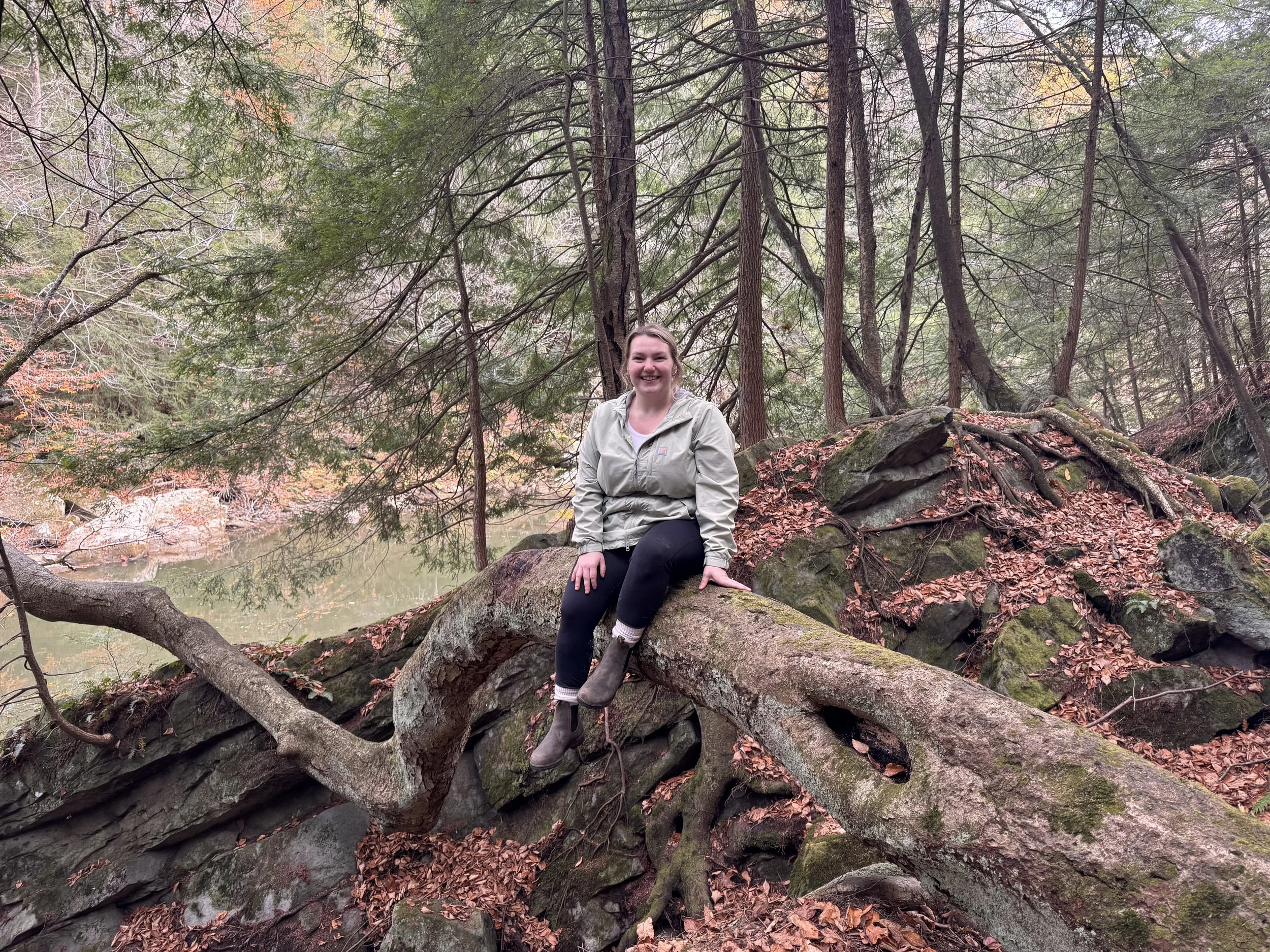 A woman smiling and wearing a beige jacket, sitting on a large rock among trees in a forested area with fallen leaves and a body of water in the background.