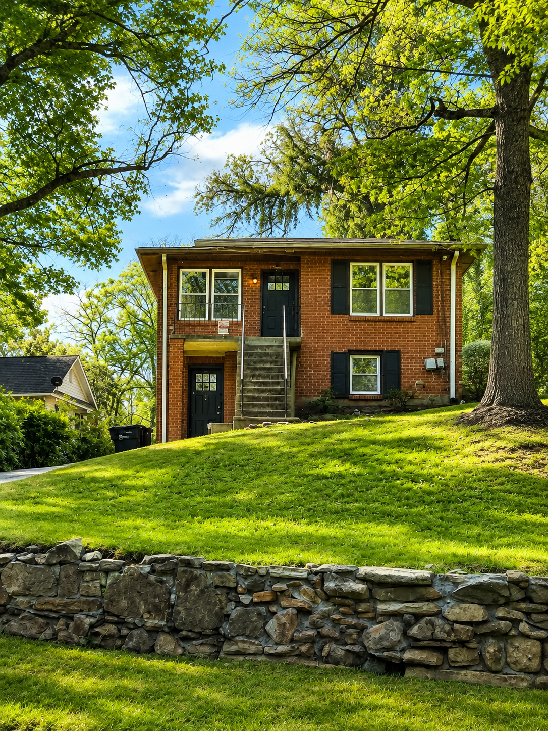 A two-story brick house with black shutters and a front staircase, surrounded by green grass and tall trees with leafy branches.
