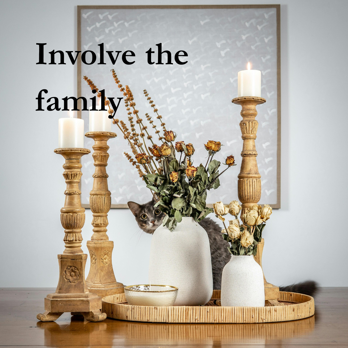 Decorative wood tray with two white vases with dried flowers, three lit candles on wooden candle holders, and a small bowl on a wooden table. Cat peeks from behind the smaller vase.  text says "Involve the family".