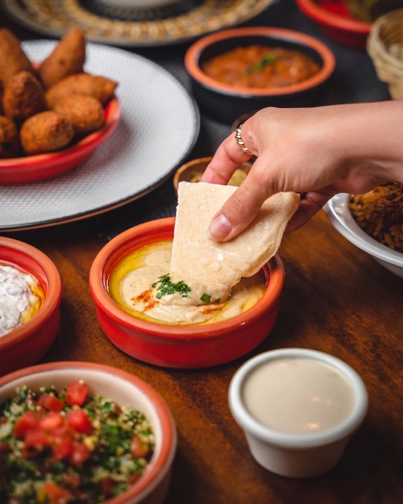 Persona mojando un trozo de pan en un plato con salsa o hummus en una mesa con otros platos de comida mexicana.