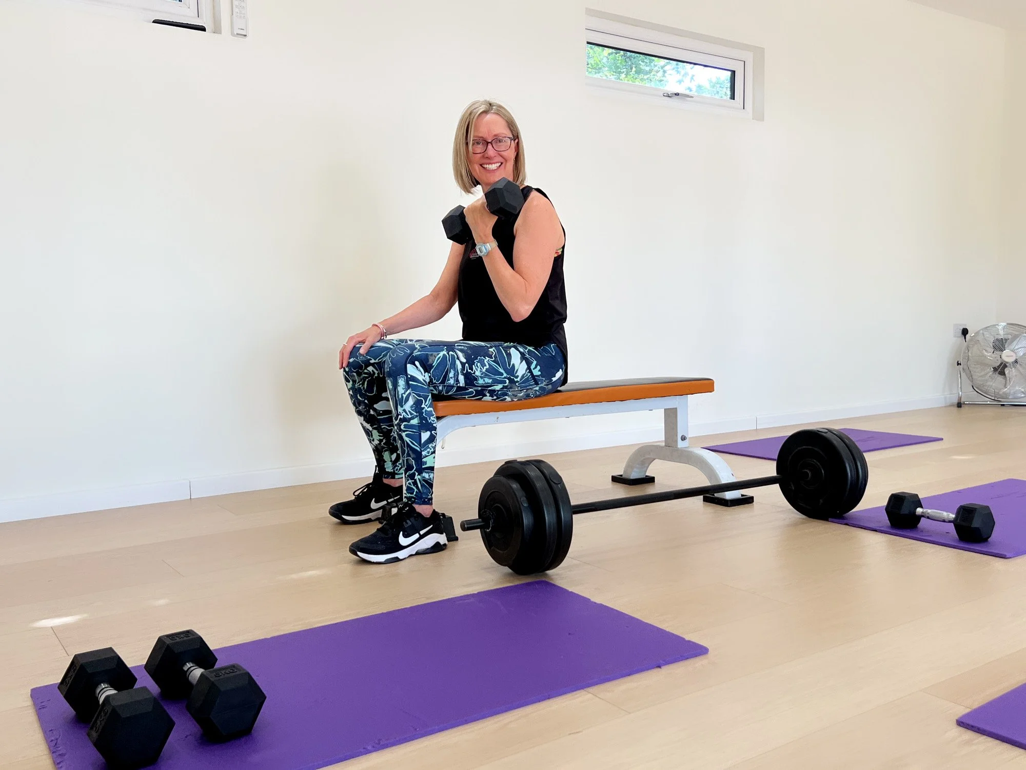 A woman with glasses smiling while sitting on a bench in a fitness room, holding a dumbbell in her right hand. She is dressed in workout clothes with patterned leggings and black sneakers. The room has a purple yoga mat, two black dumbbells, a barbell, and a purple exercise mat on the light wood floor. A fan is on the right, and two small windows are visible on the wall behind her.