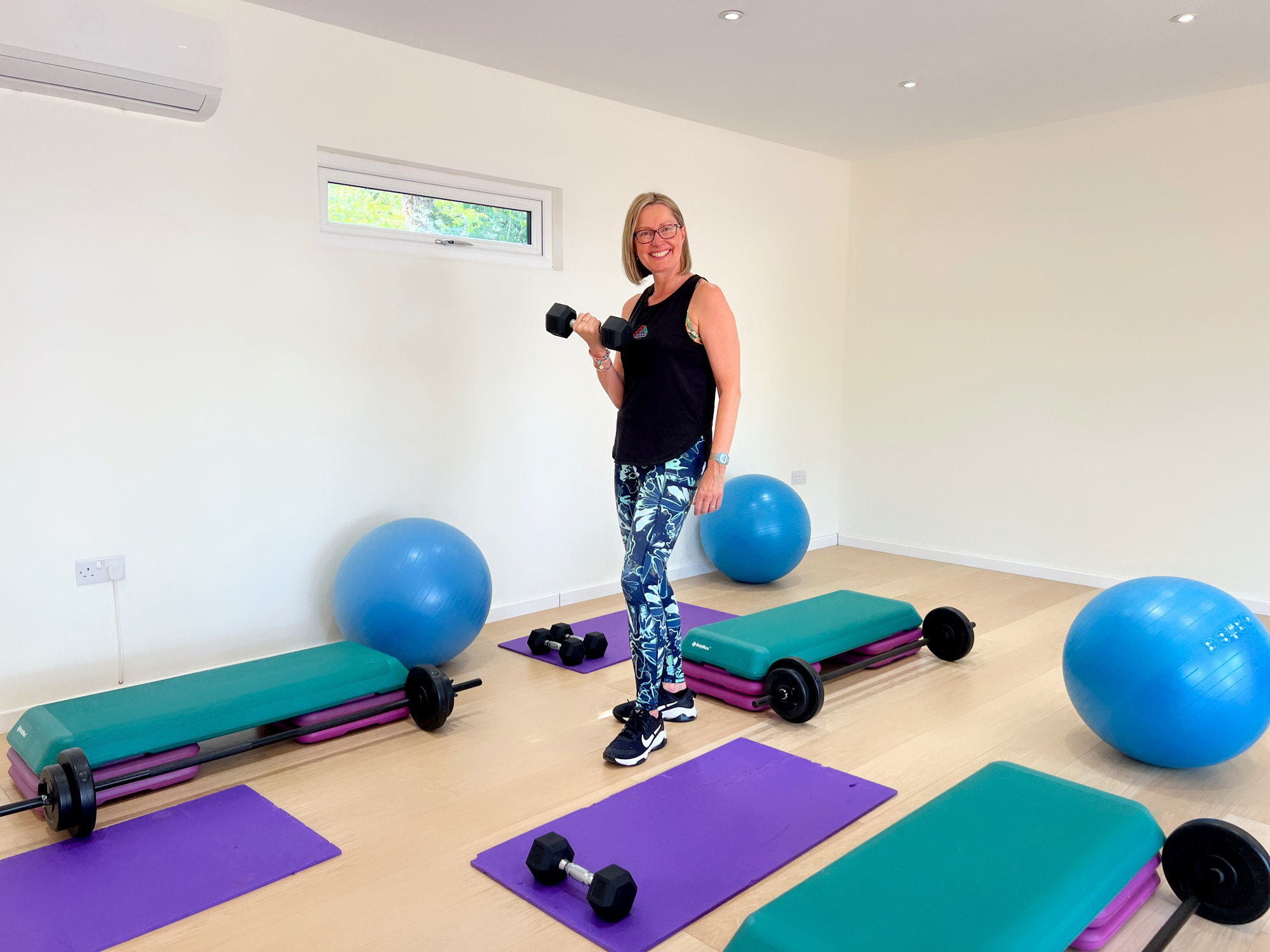 A woman in workout attire holding a dumbbell in a home gym with exercise mats, fitness balls, dumbbells, and step platforms.