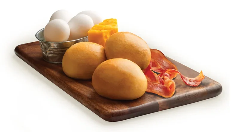 Three Kolaches, three eggs, sliced cheddar cheese, bacon strips on a wooden cutting board