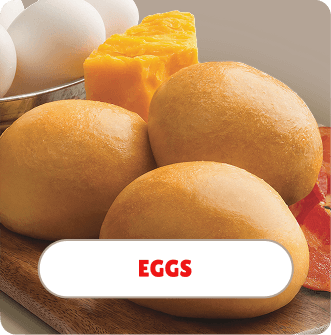 Three bread rolls, a block of yellow cheese, and a cluster of eggs on a wooden surface.